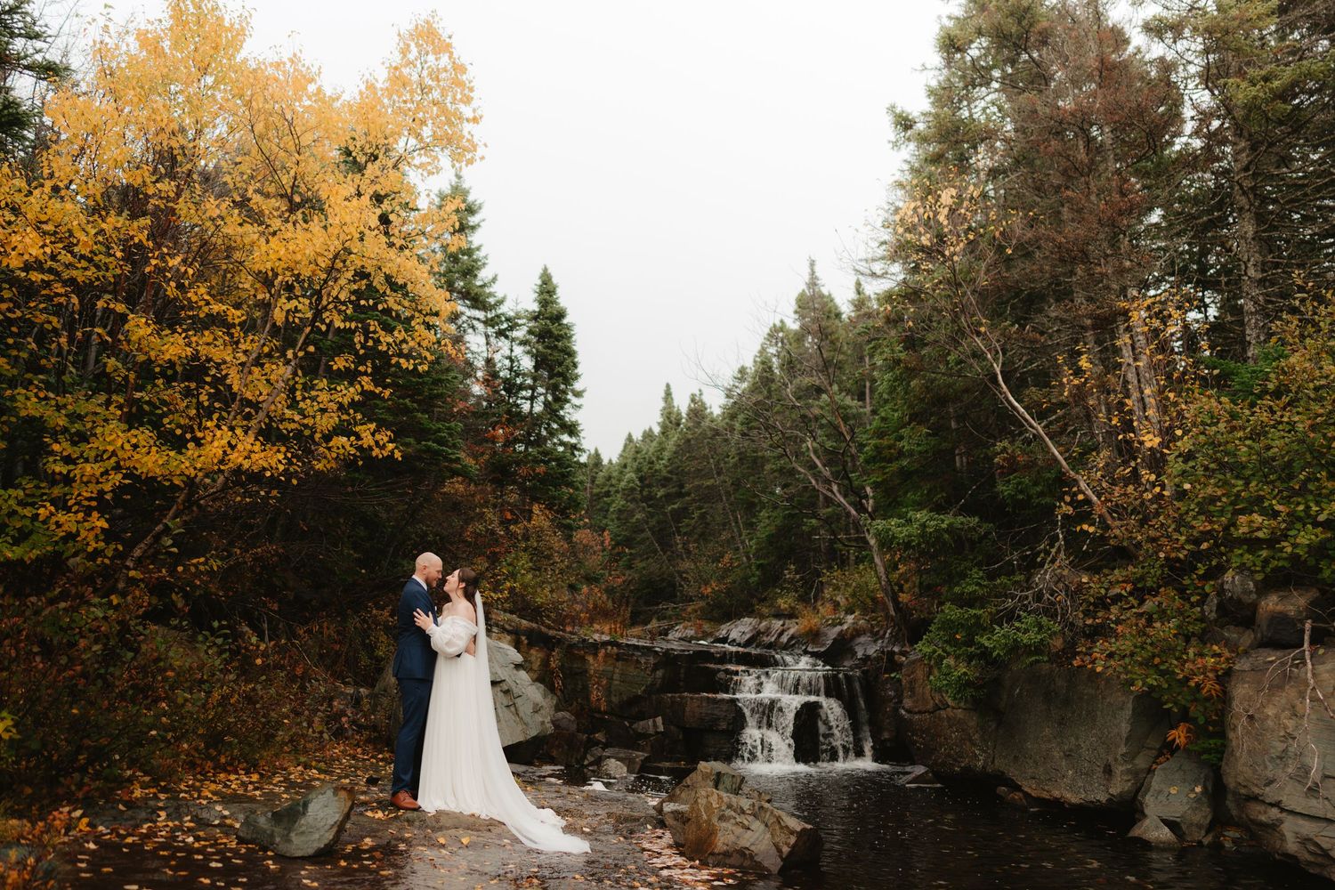 A couple shares a romantic moment by a waterfall surrounded by vibrant autumn foliage in a forest setting.
