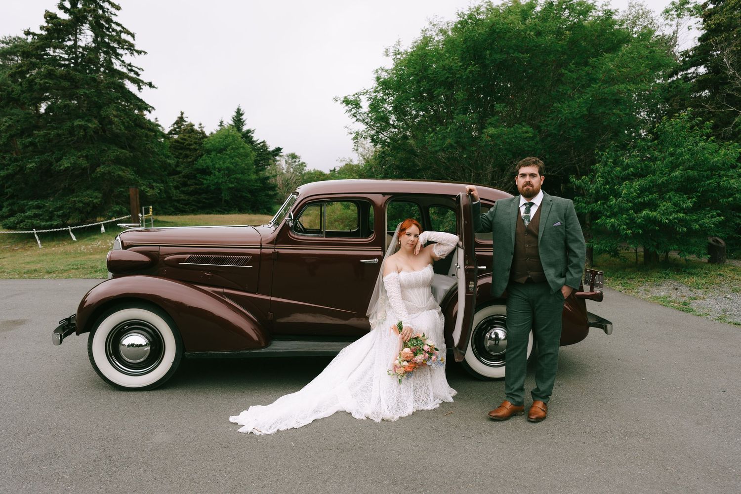 Bride and groom posing beside a vintage brown classic car outdoors, surrounded by lush green trees.