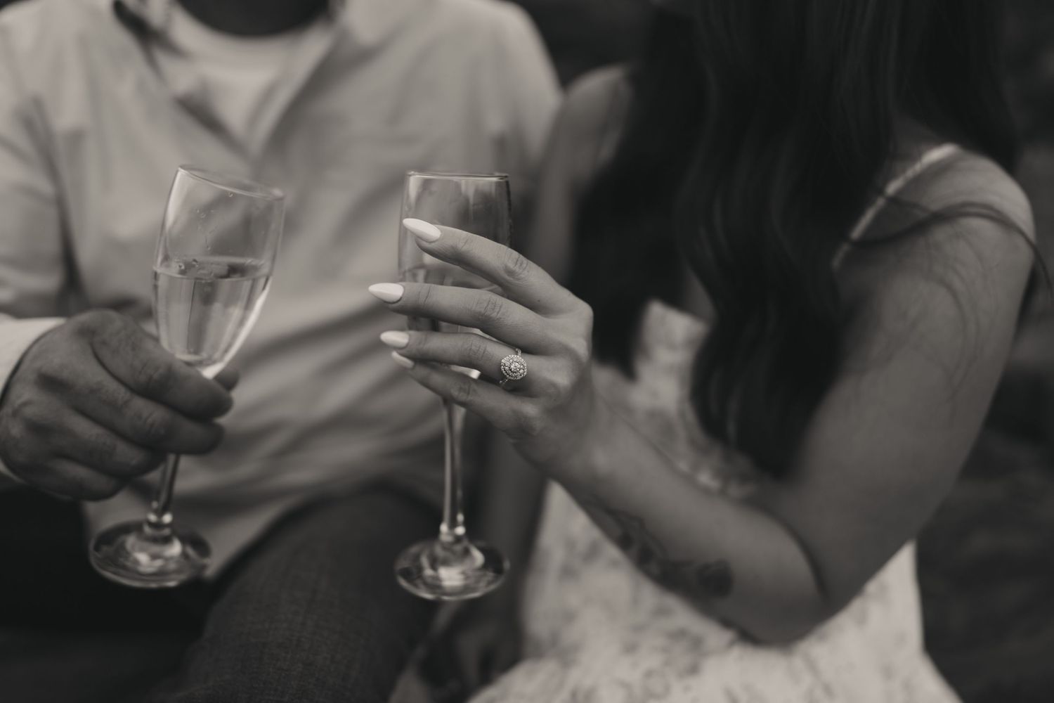 Black and white photo of couple holding champagne glasses, woman wearing engagement ring, celebrating romantic moment.