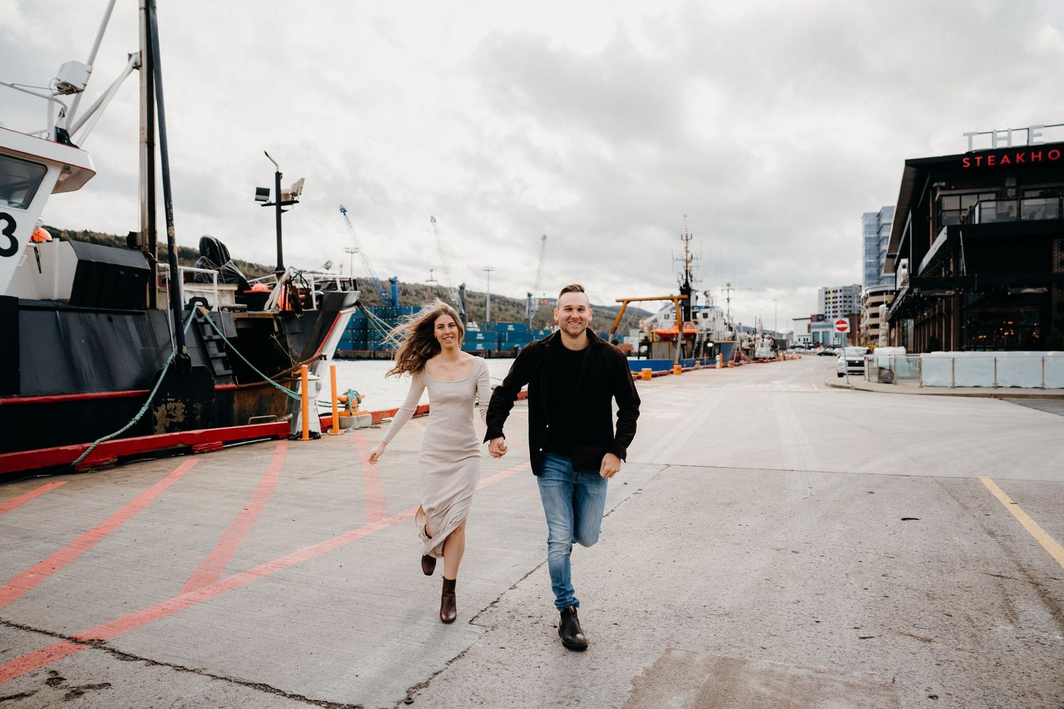 A couple walks hand in hand along an industrial waterfront dock.