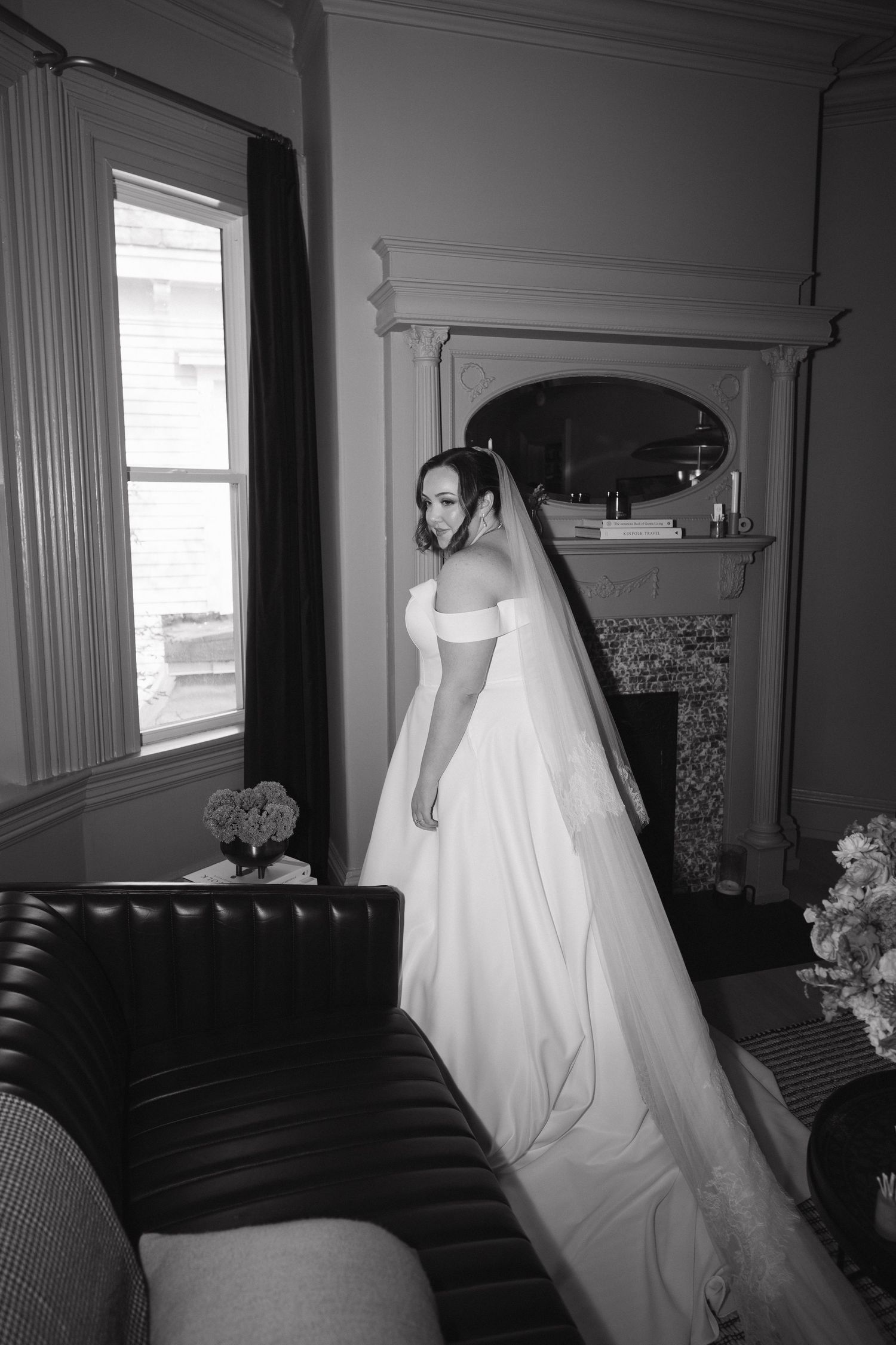 White wedding dress hanging in a classic room with dark furniture and window light.