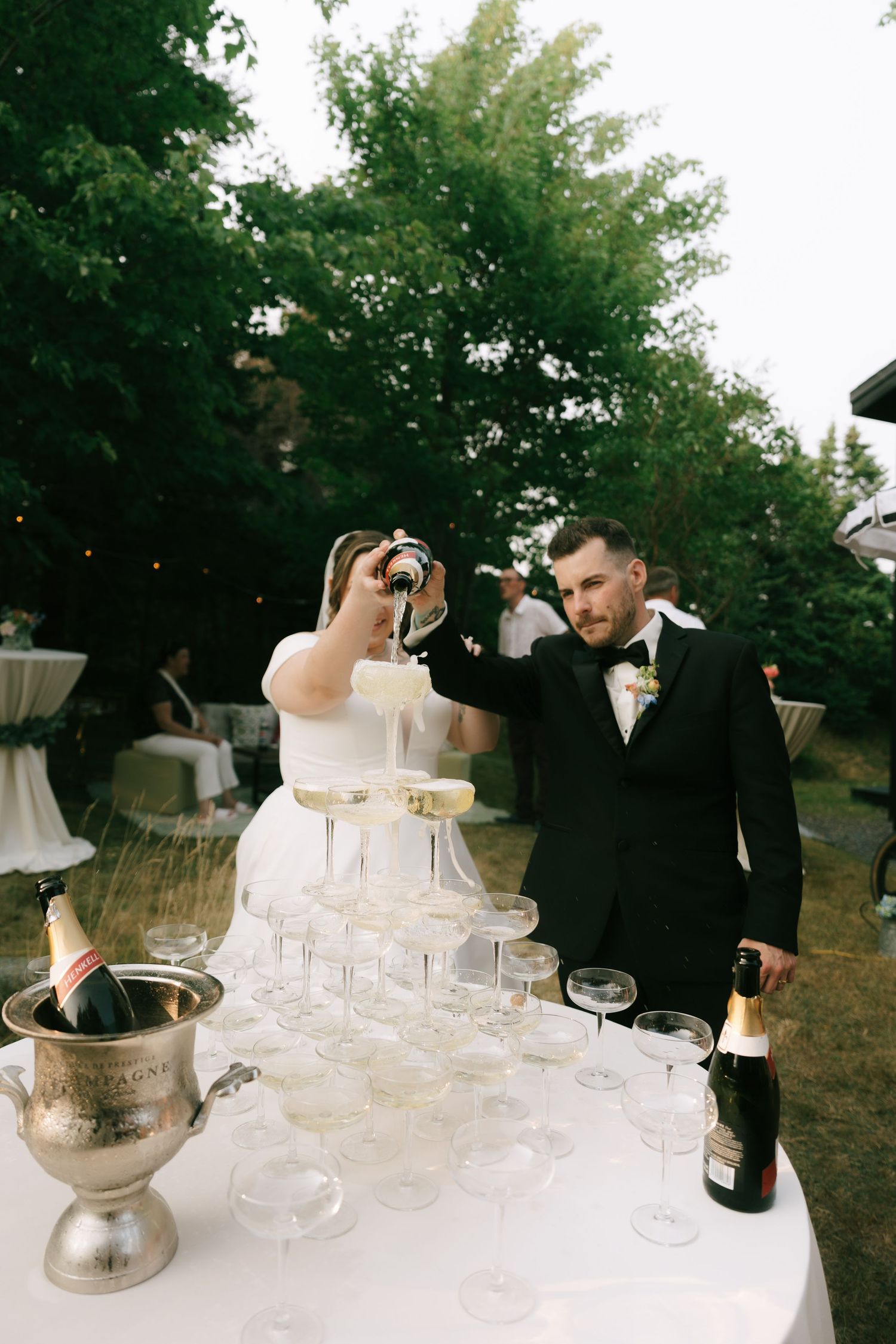 Champagne tower being filled at an outdoor celebration with trees in the background.