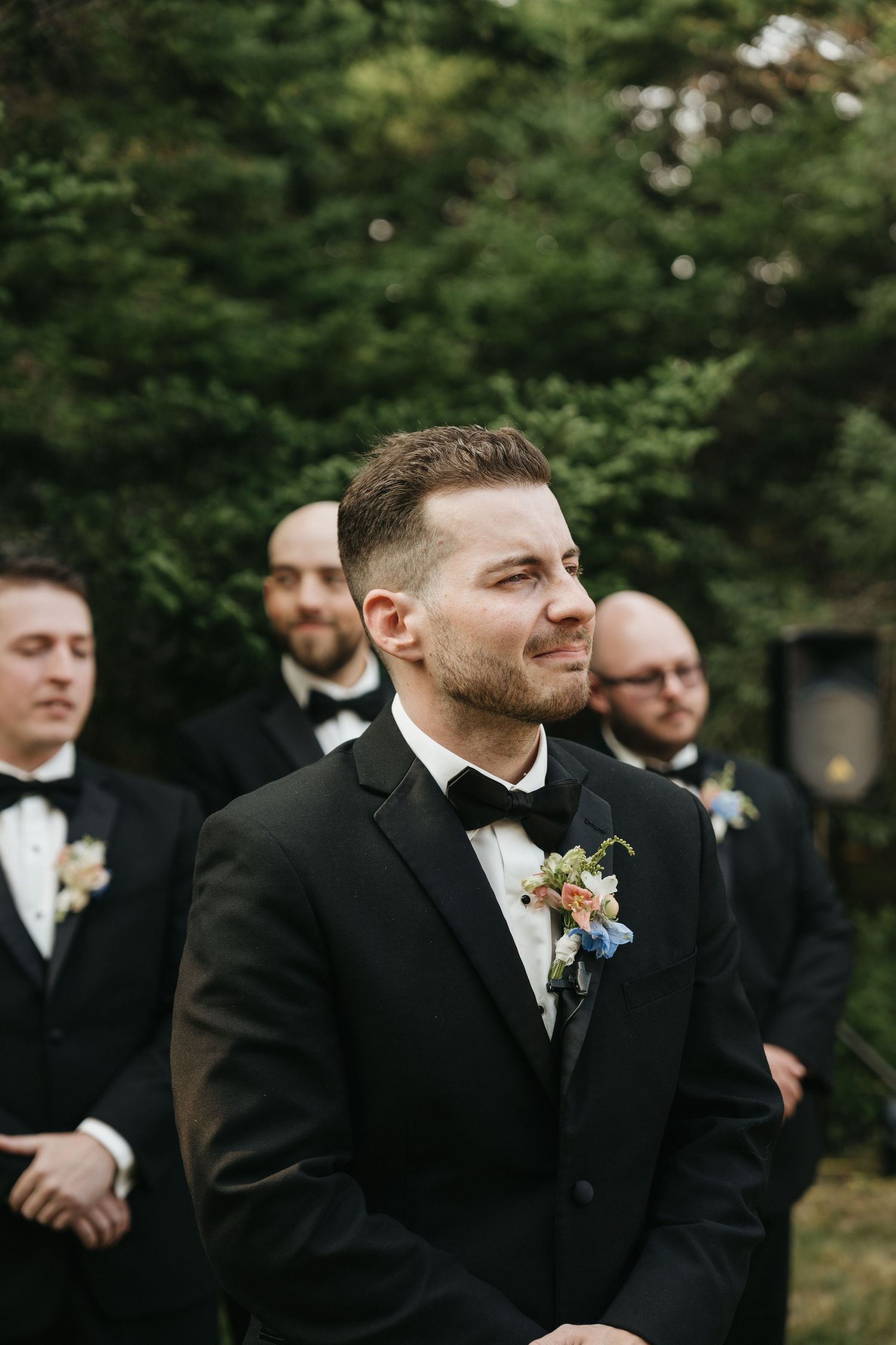 Wedding party in black suits and boutonnieres standing in front of evergreen trees.