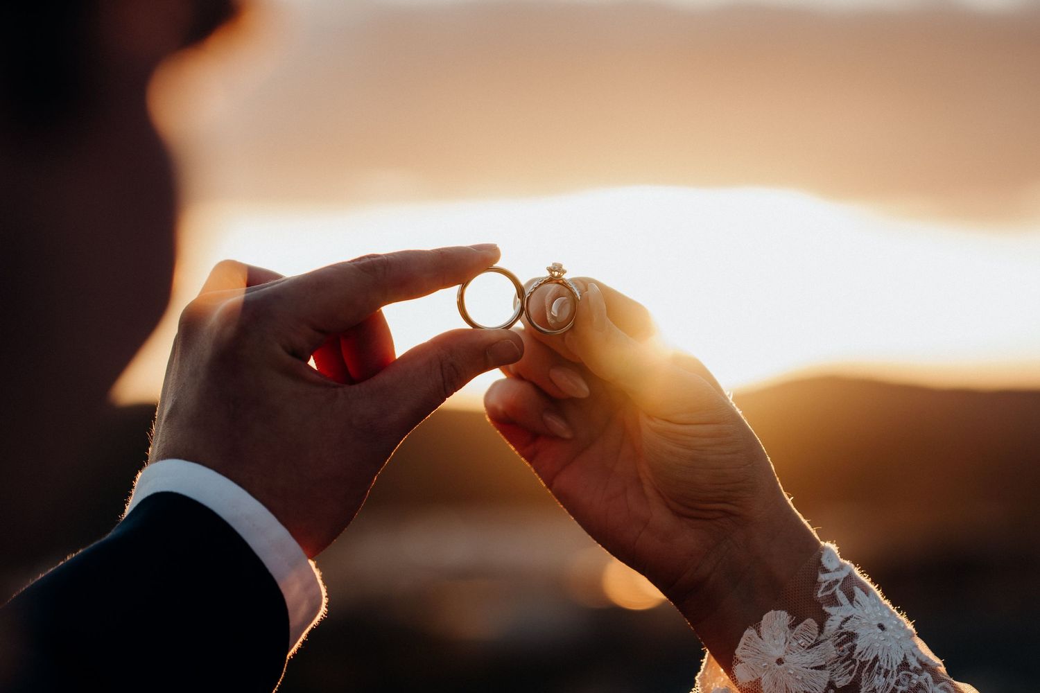 Close-up of hands holding wedding rings against golden sunset light.