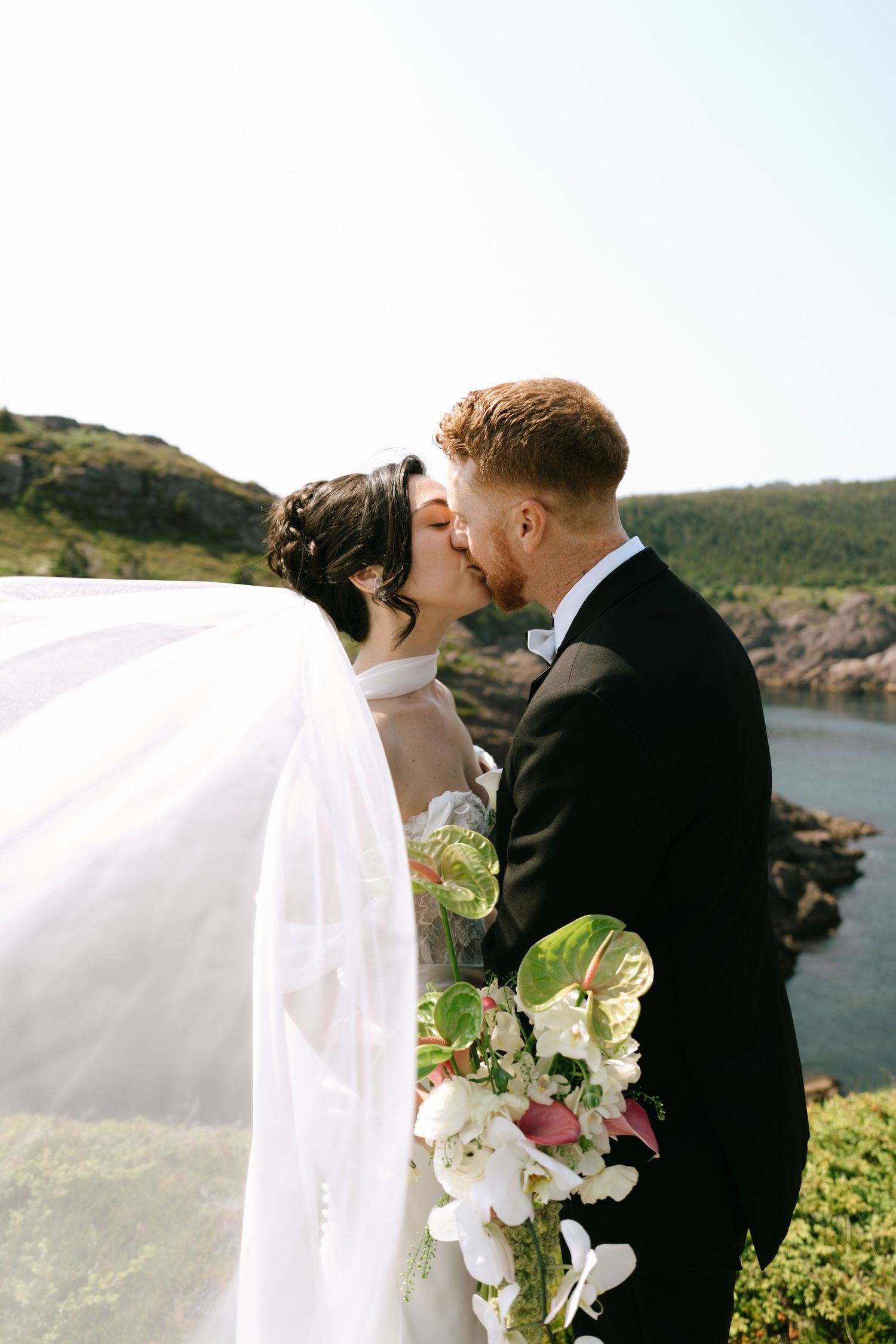 Bride and groom share a kiss outdoors with veil flowing, holding white floral bouquet against scenic coastal cliffs.