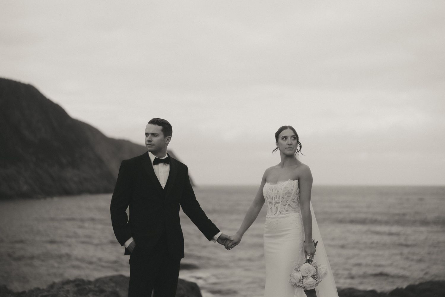 Black and white photo of a bride and groom holding hands by the ocean cliffs on their wedding day.