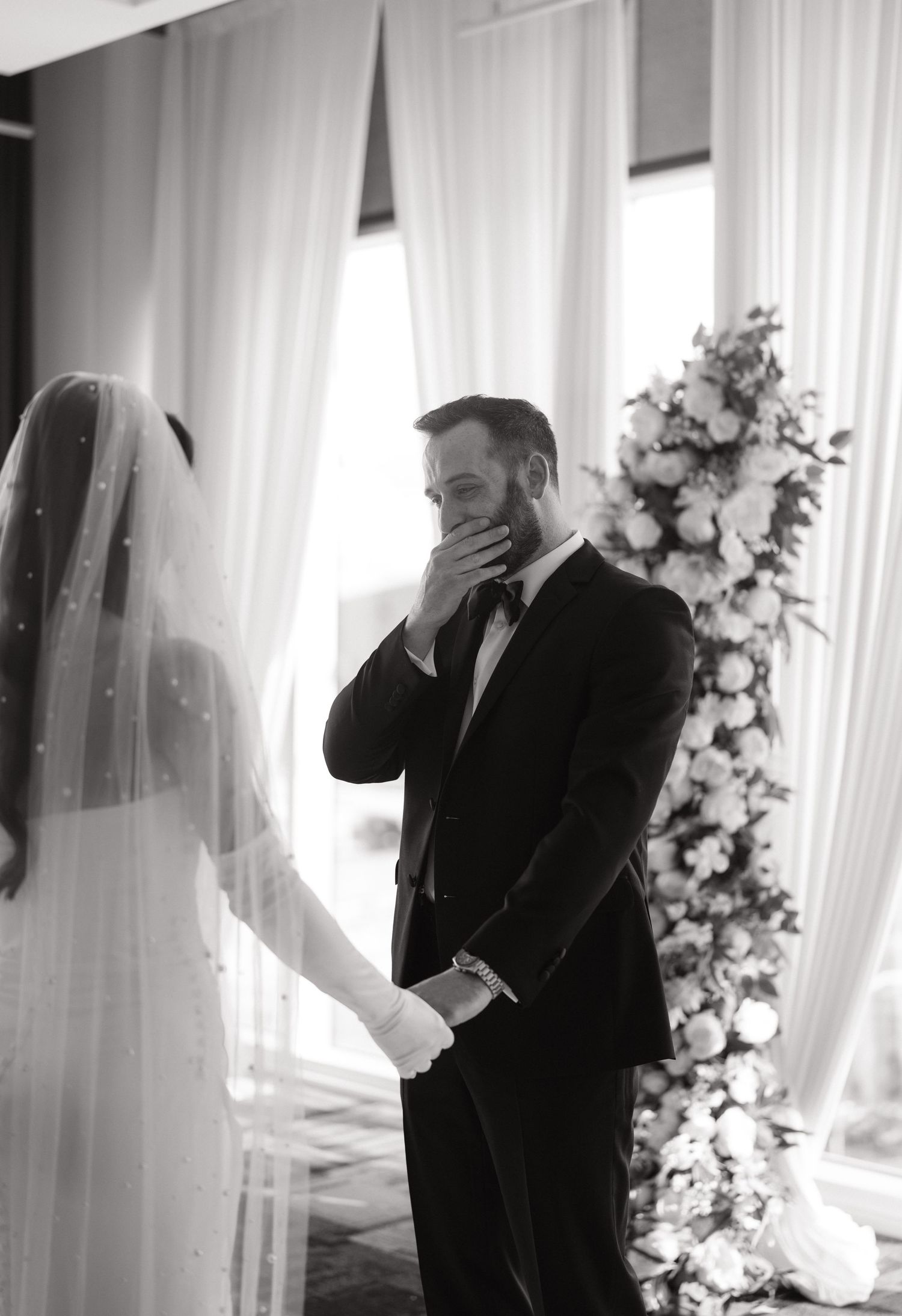 Black and white photo of emotional groom covering mouth as bride approaches at elegant wedding ceremony with floral arch.
