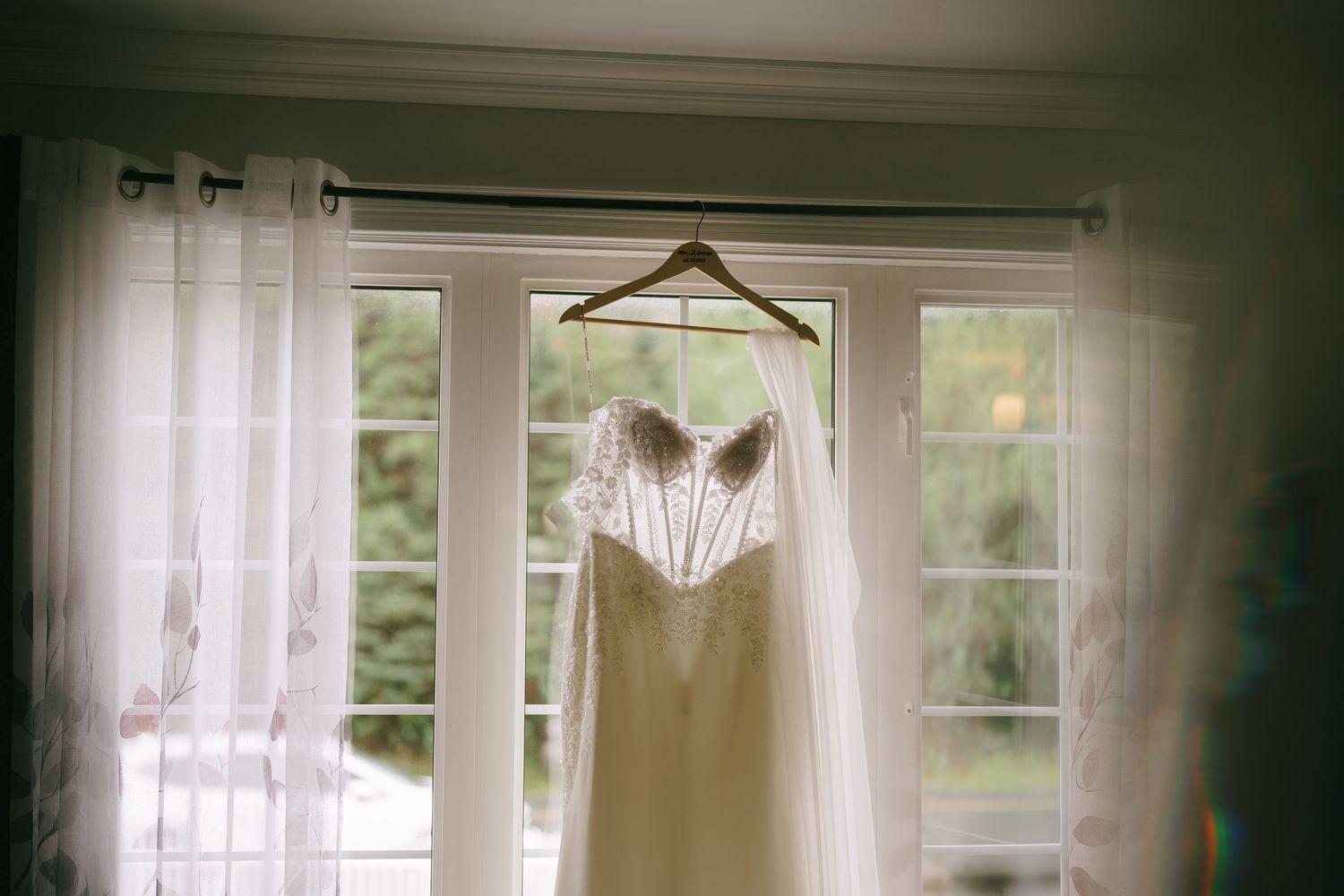 Elegant wedding dress with lace bodice hanging on wooden hanger in front of bright window with sheer curtains.