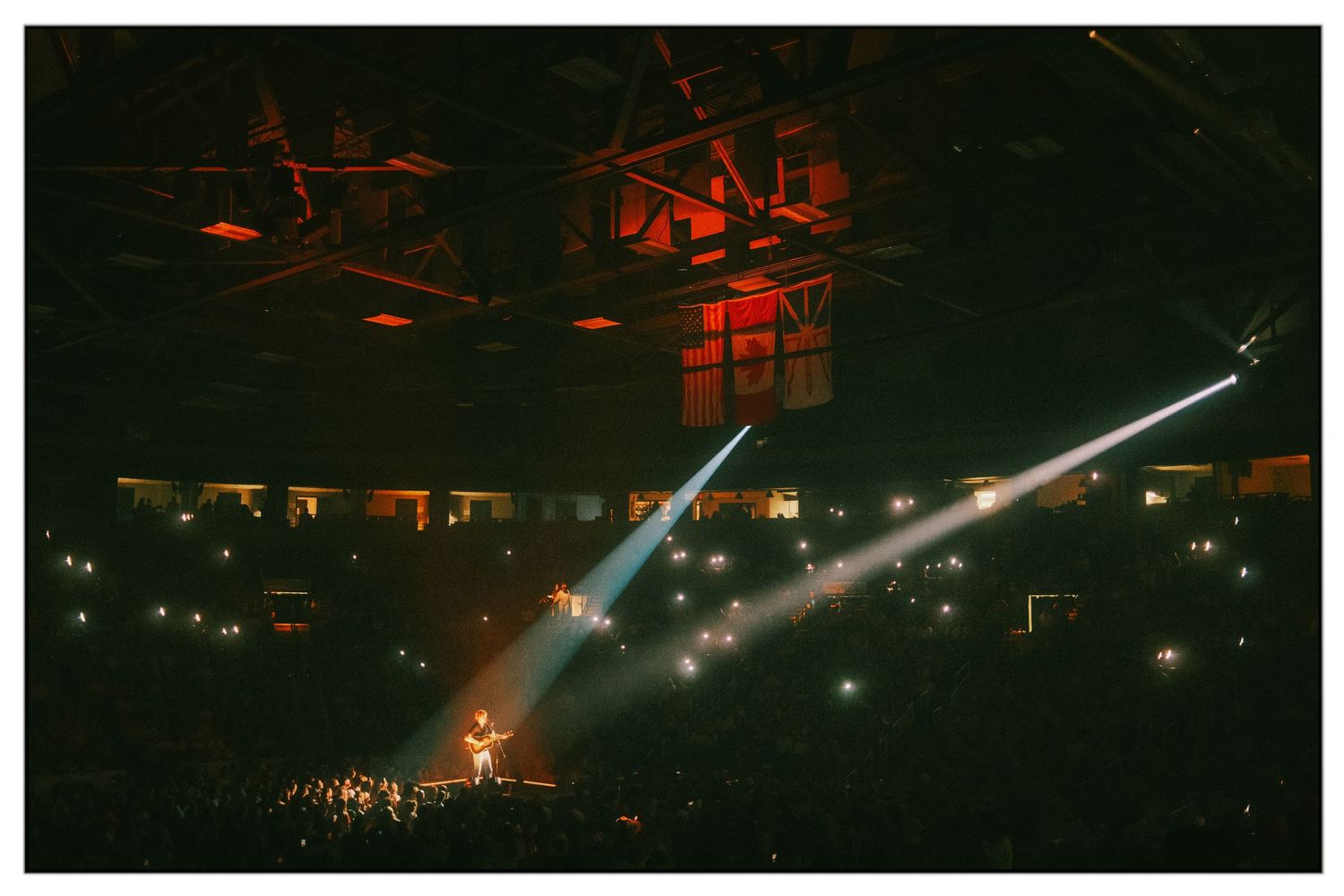Bright white spotlights illuminate a dark concert venue with red lighting effects creating an atmospheric ambiance.