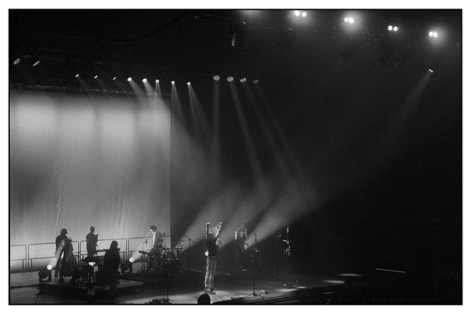 Dramatic concert stage illuminated with bright spotlights and fog creating silhouettes of performers during live performance.