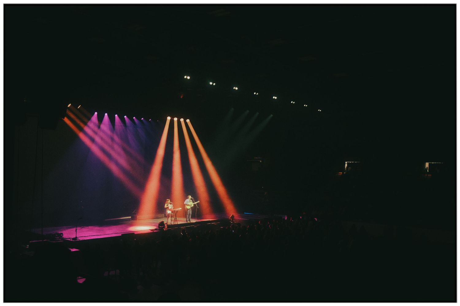 Colorful stage lighting creates vibrant pink and orange beams across a dark performance venue.