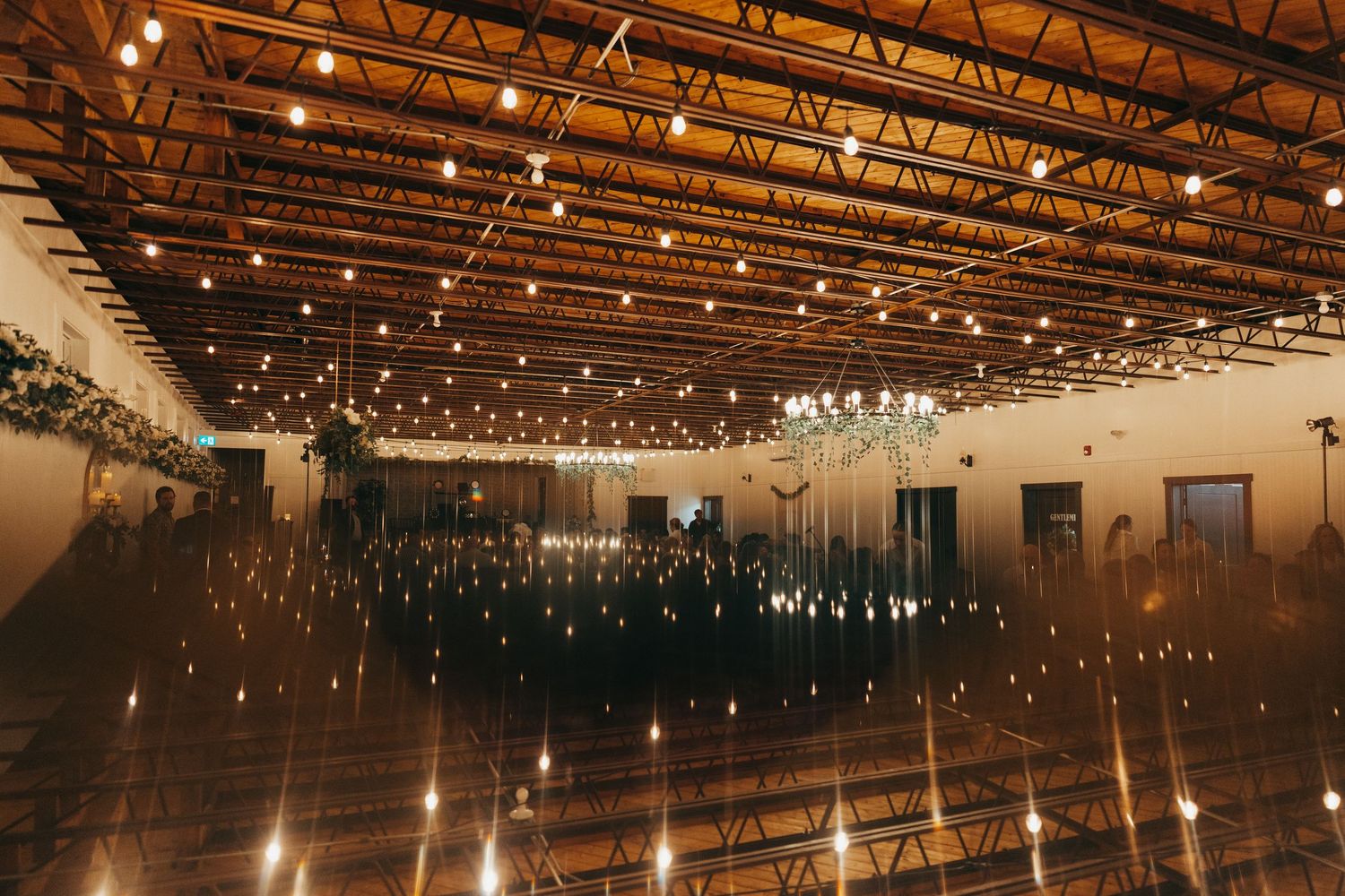 Rustic indoor wedding venue with exposed wooden beams and twinkling string lights creating a warm, romantic atmosphere.