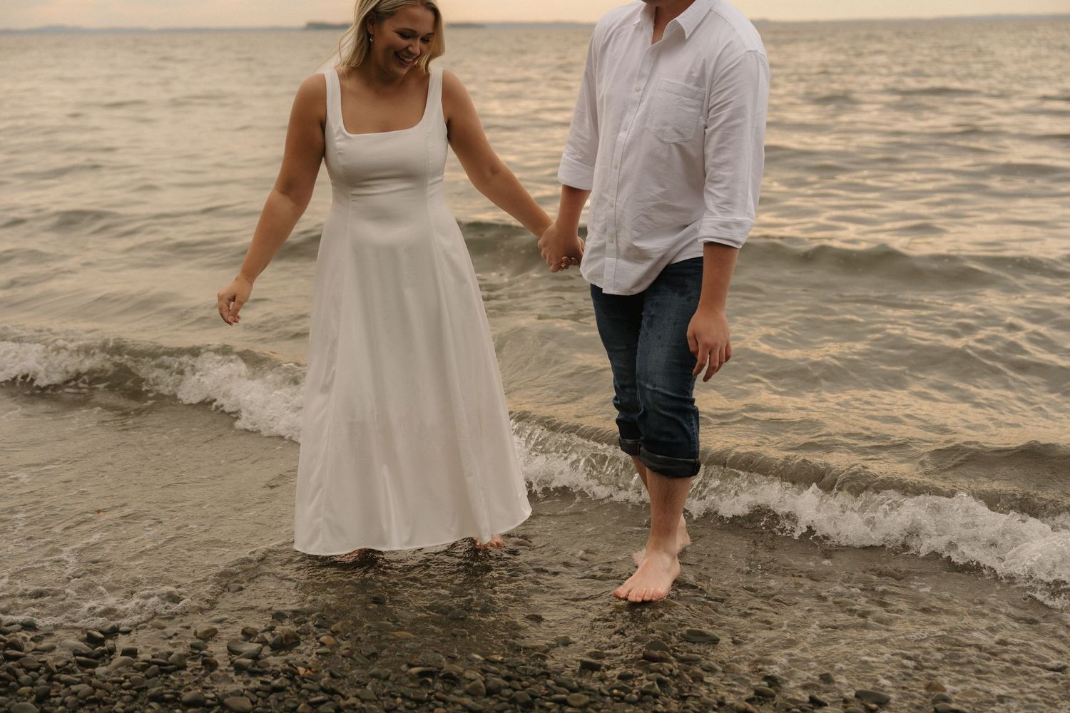 Couple strolling together through shallow ocean water at sunset with white dress flowing in the breeze.