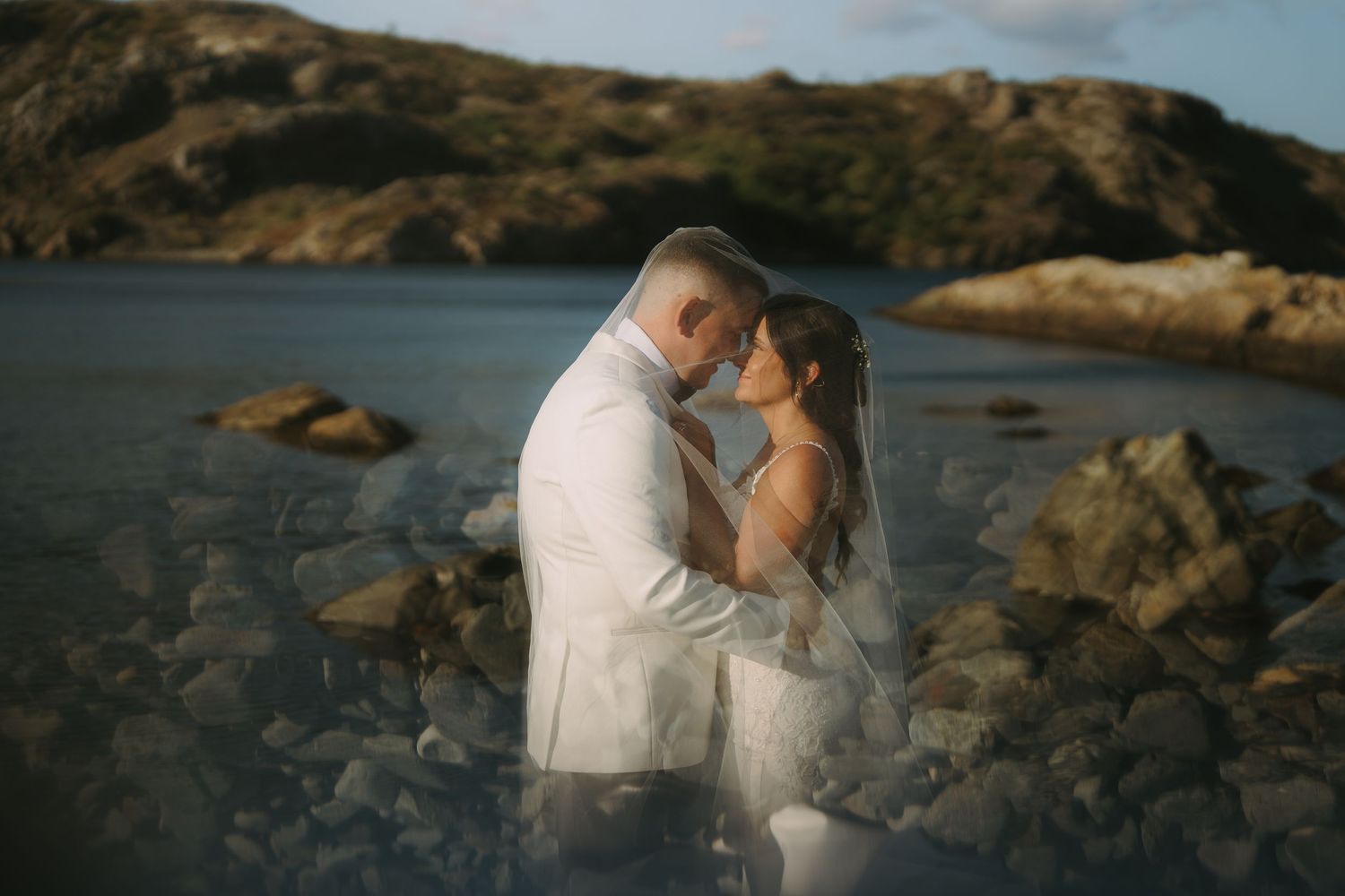 Two people in wedding attire share an intimate moment on rocky coastal cliffs at sunset.