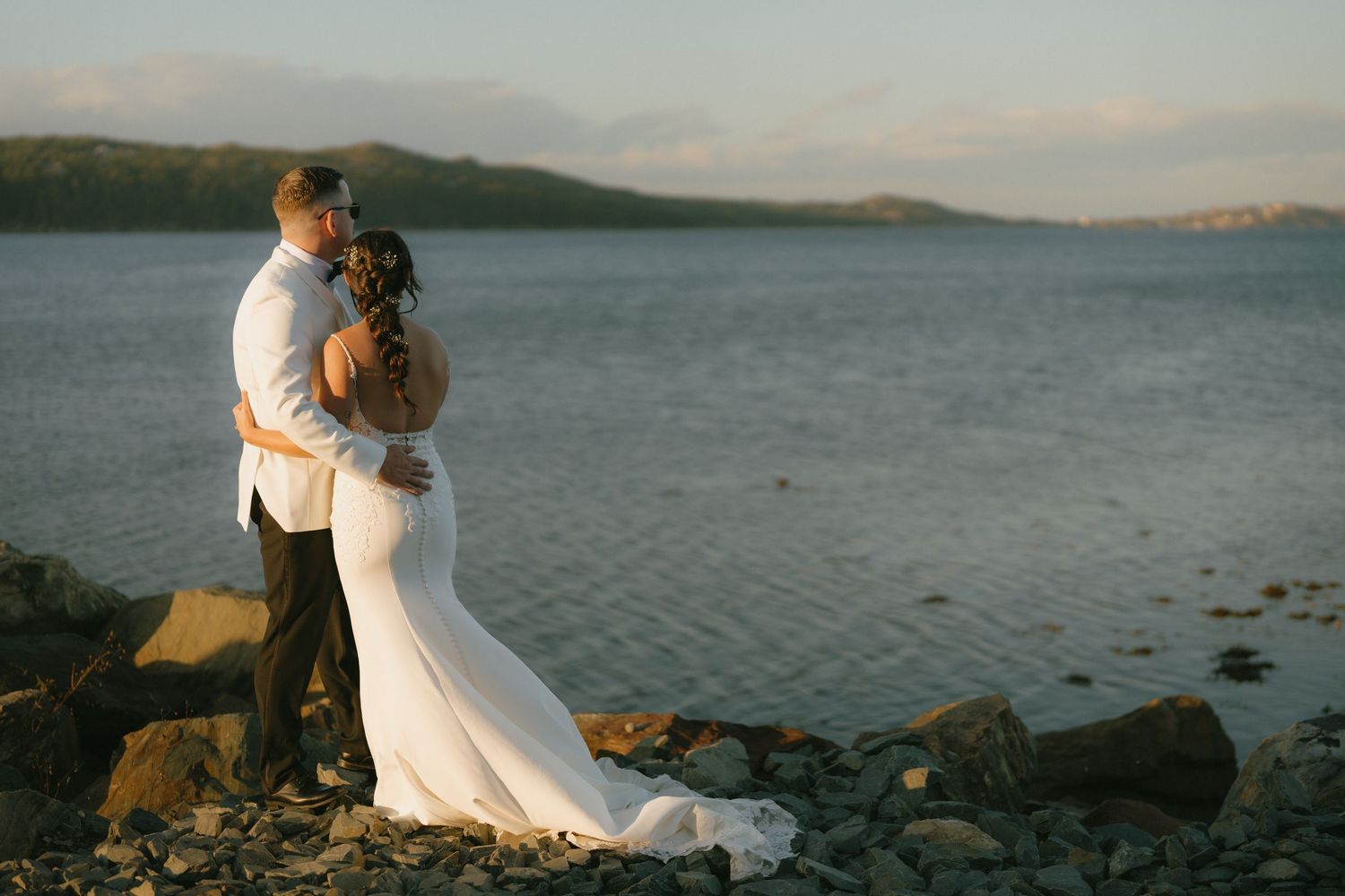 A romantic couple embraces on a rocky shoreline at sunset with calm ocean waters and mountains in the background.