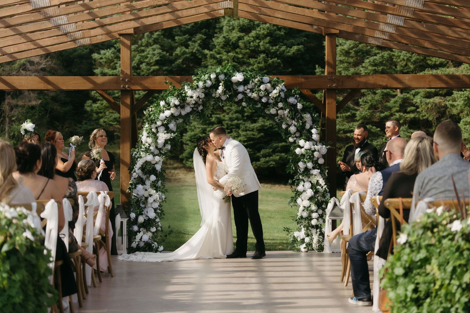 A romantic outdoor wedding ceremony takes place under a rustic wooden pergola adorned with white floral arrangements.