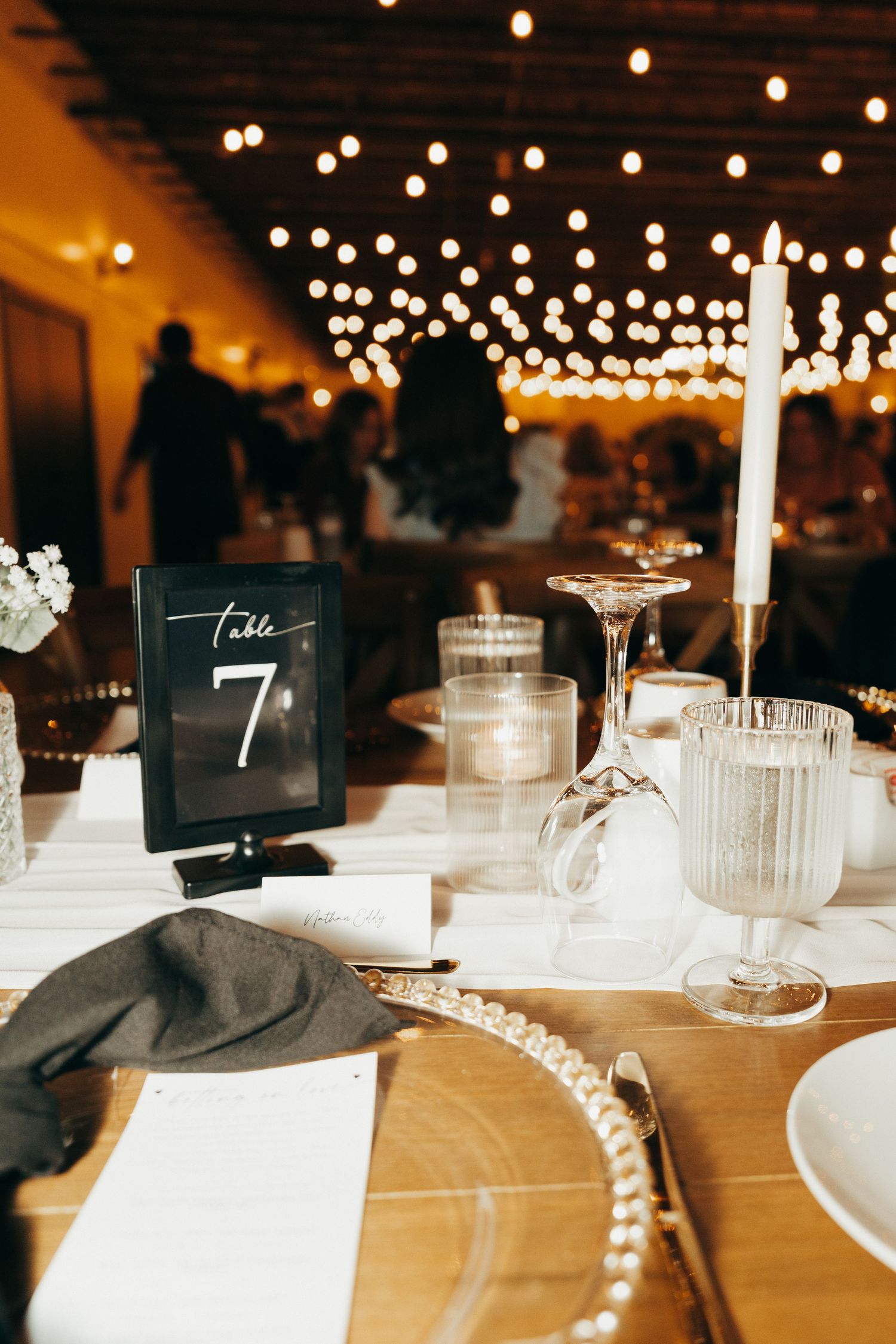 Wedding reception table settings with black table numbers and glowing string lights overhead create an elegant atmosphere.