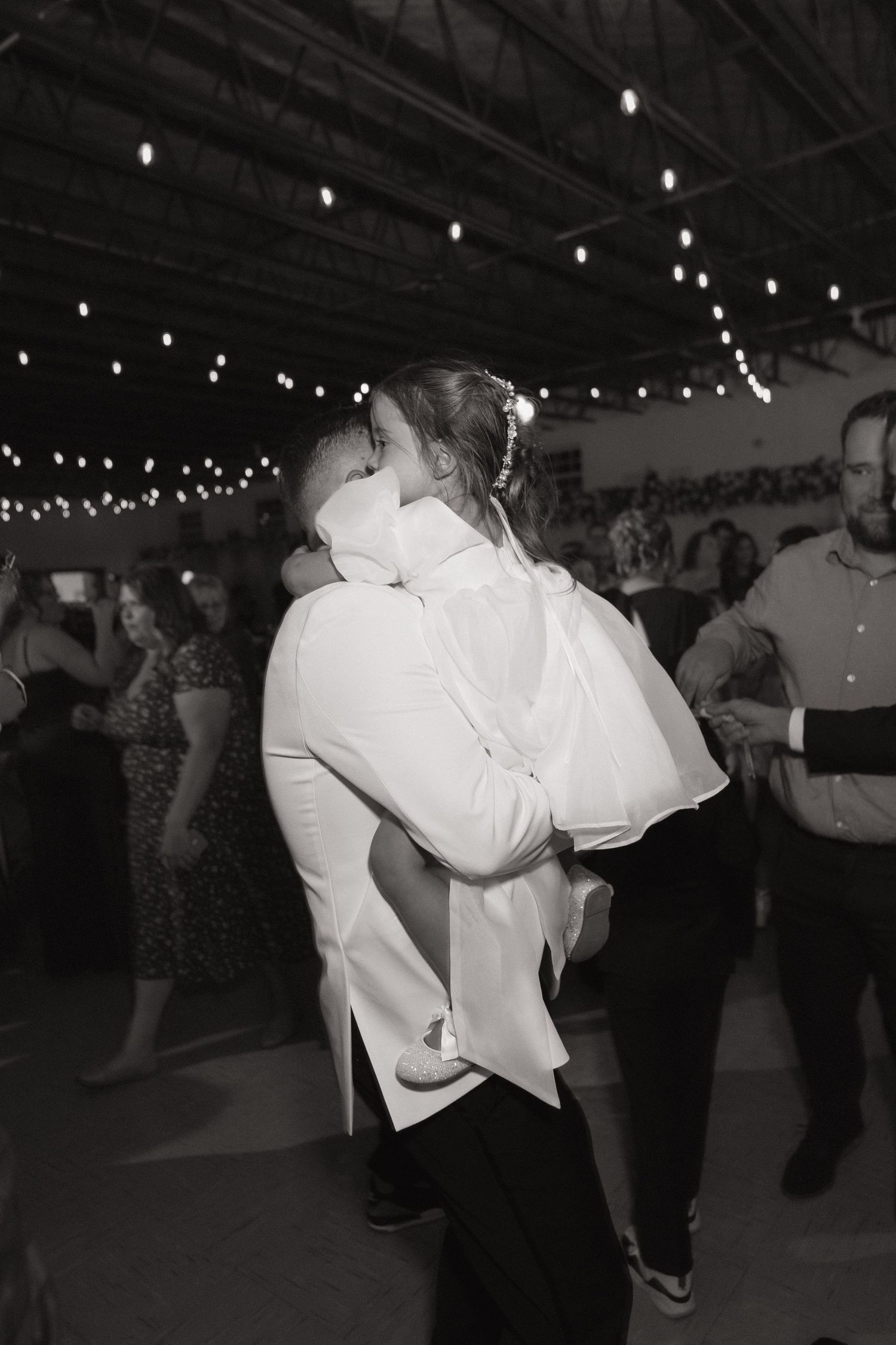 A couple in white attire dances together at a dimly lit indoor venue with string lights overhead.