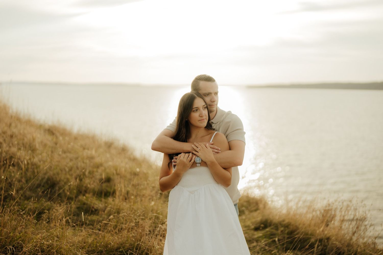 A romantic couple embraces on a sunlit hillside overlooking the ocean during a golden hour photoshoot.