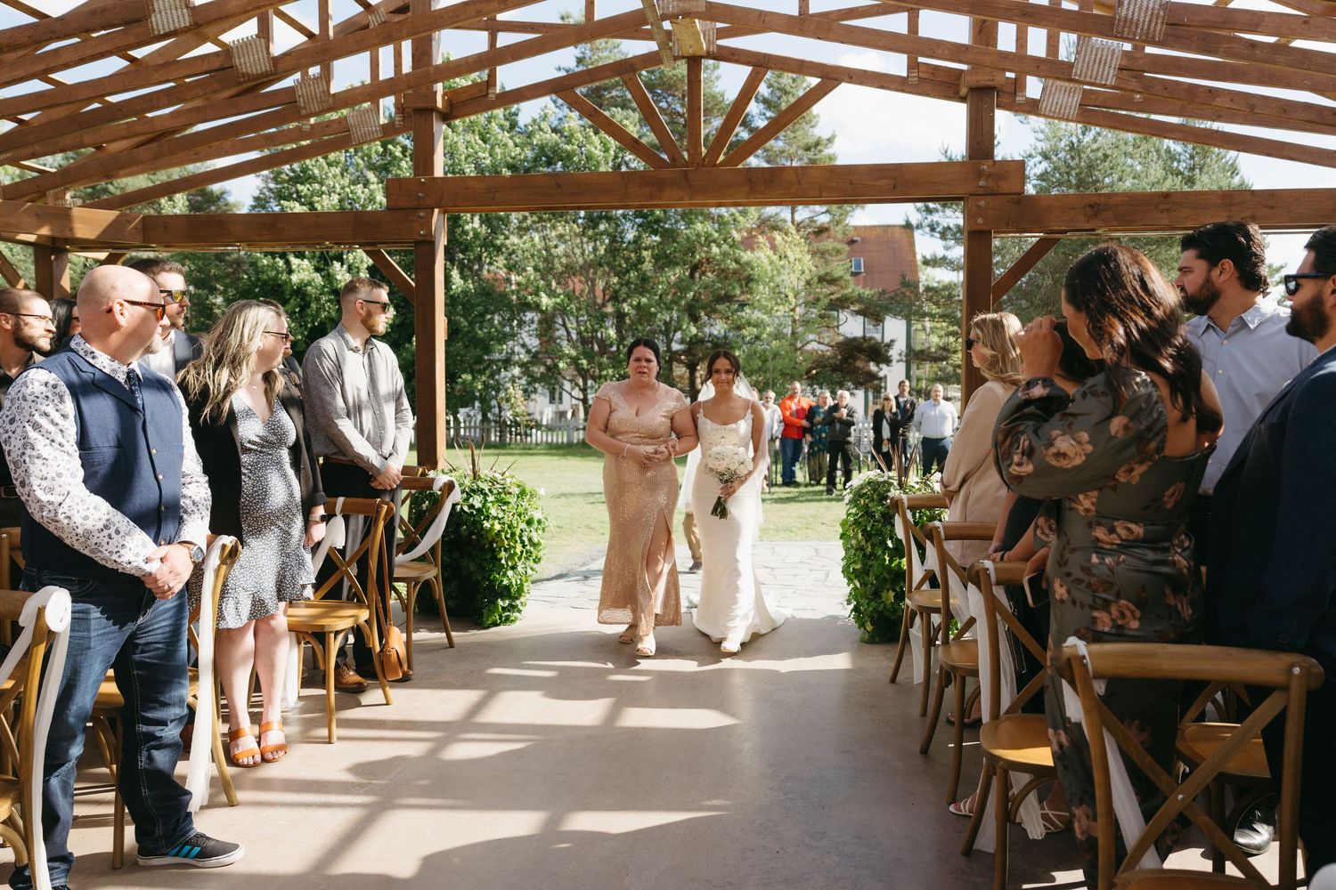 Wedding guests mingle under a rustic wooden pergola during an outdoor reception celebration.
