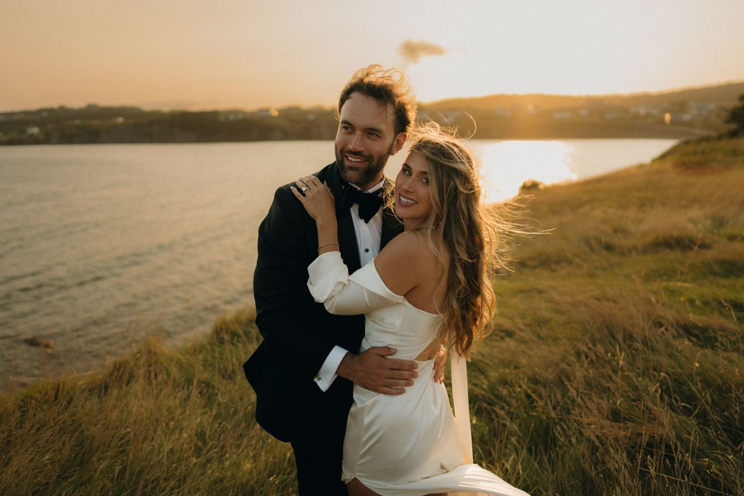 A couple embraces on a scenic coastal hillside during golden hour with rolling hills and ocean views in the background.