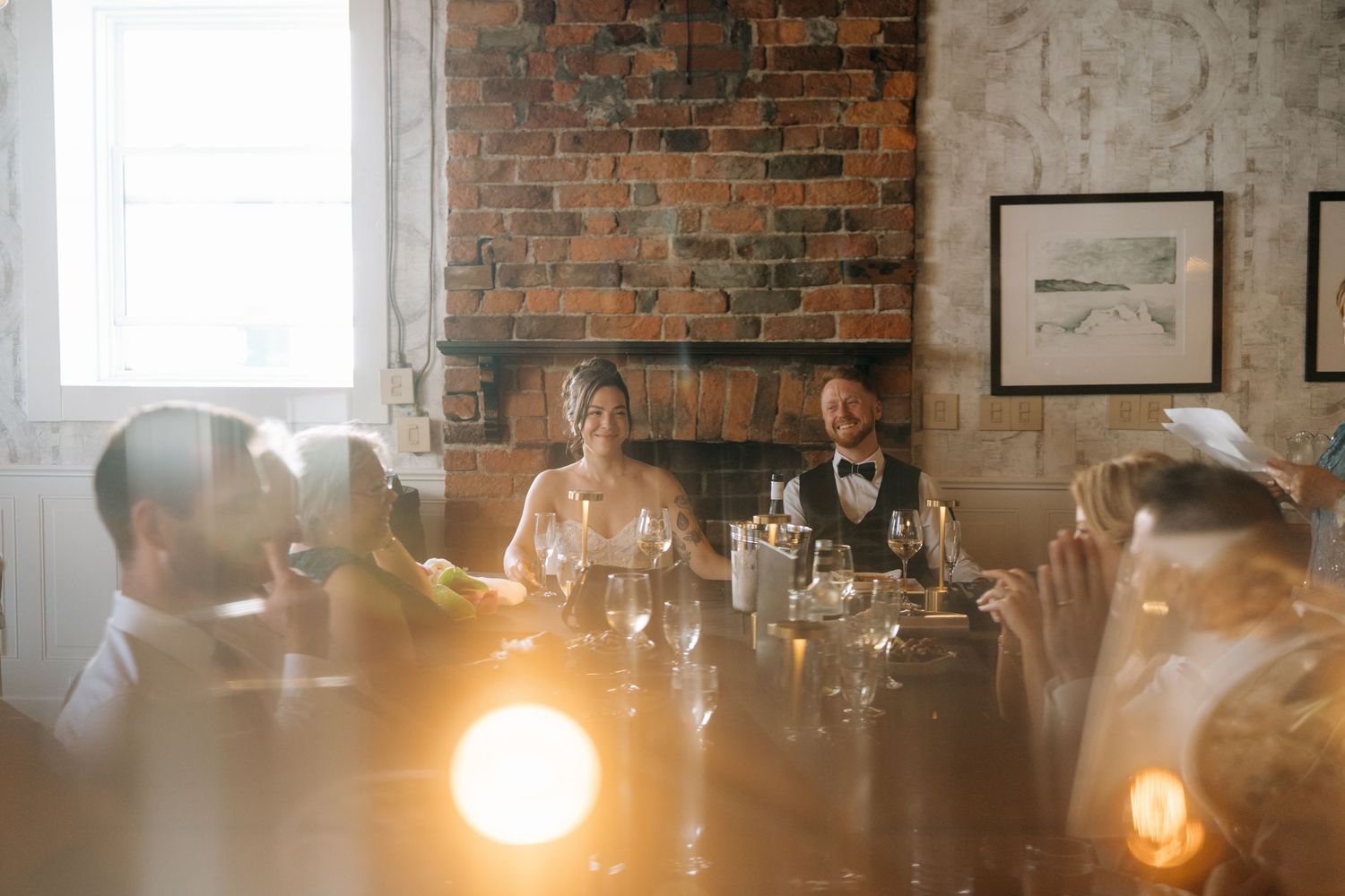 Bride and groom seated at elegant wedding reception table with exposed brick wall backdrop and warm candlelight ambiance.