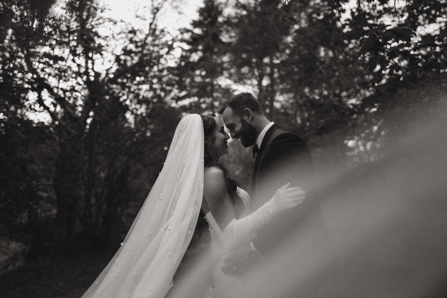 Black and white photo of a bride and groom sharing an intimate moment outdoors, surrounded by lush trees on their wedding day.