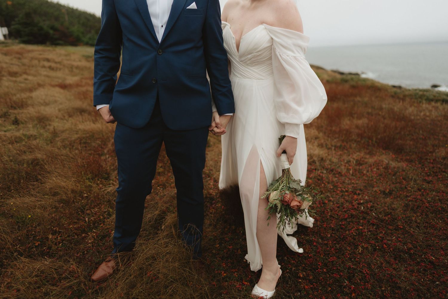 A couple in wedding attire walks together on a scenic coastal cliff with brown grass and ocean views in the background.