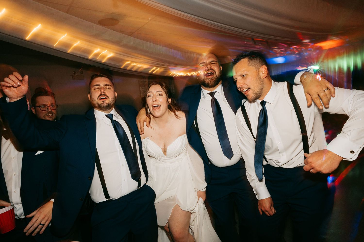 Wedding guests dance and celebrate together on a dimly lit dance floor with colorful lighting effects.