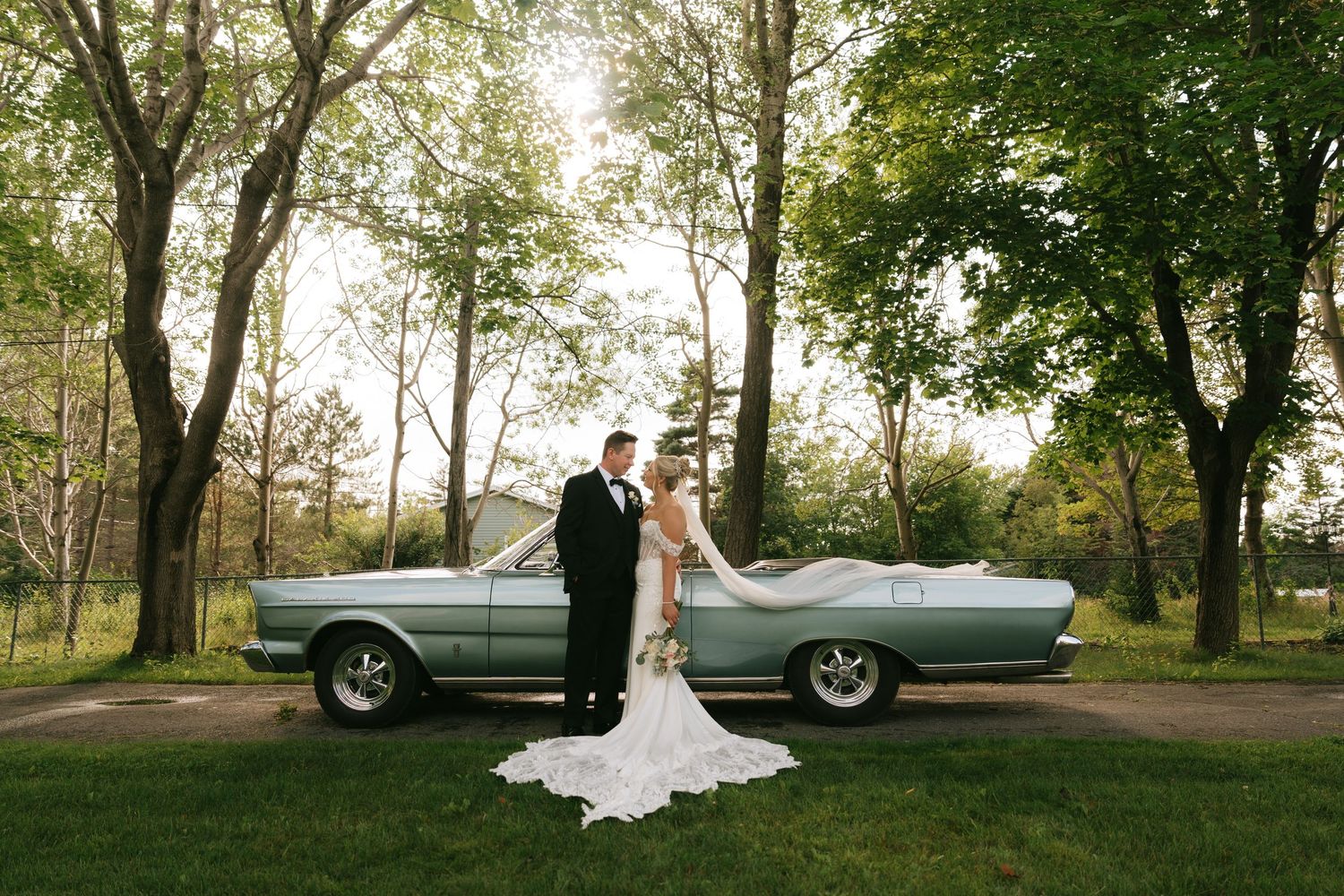 Bride and groom posing in front of a vintage light blue convertible car surrounded by lush green trees.