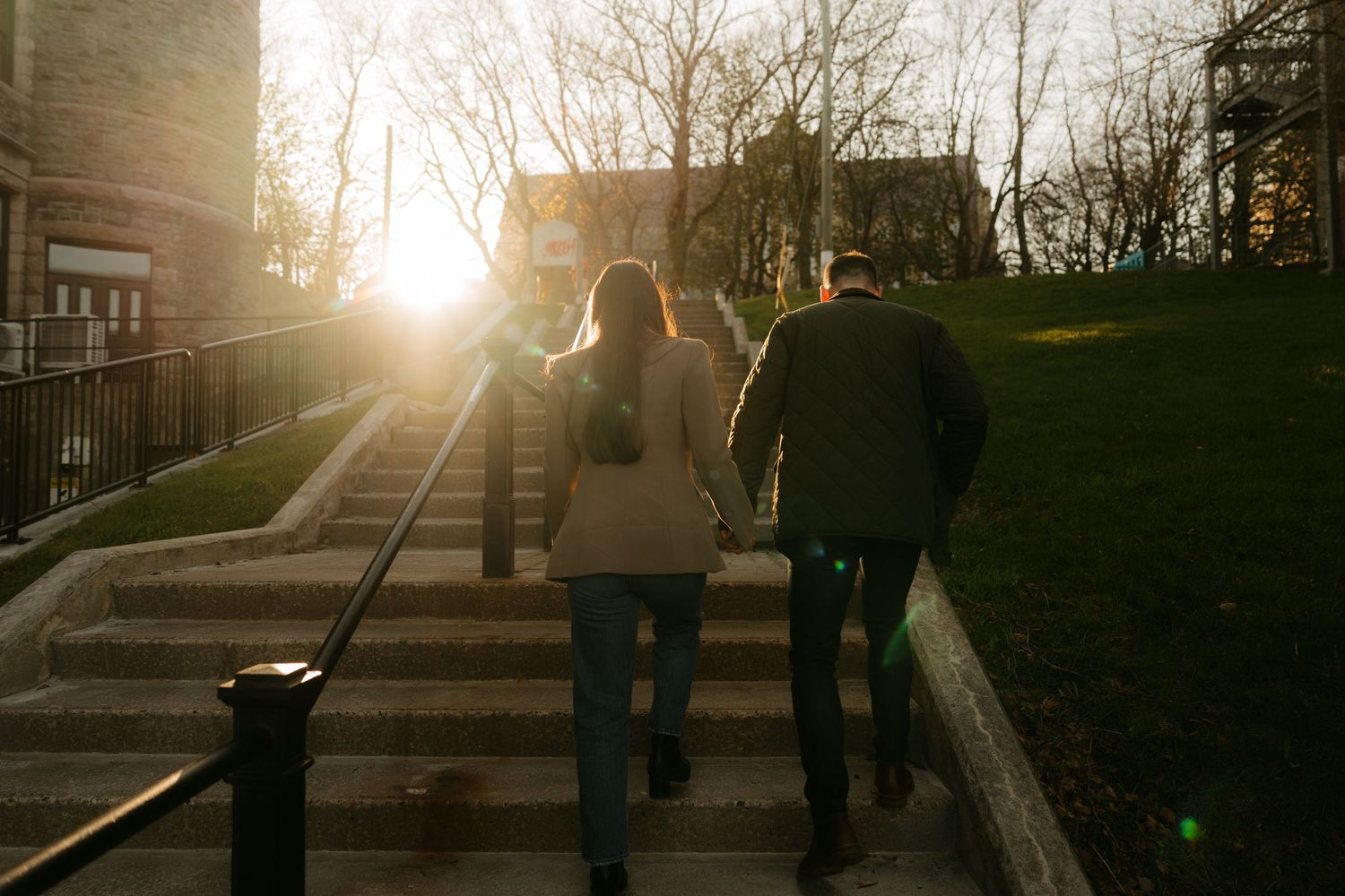 Couple walking up outdoor stairs toward a glowing sunset near a historic brick building.
