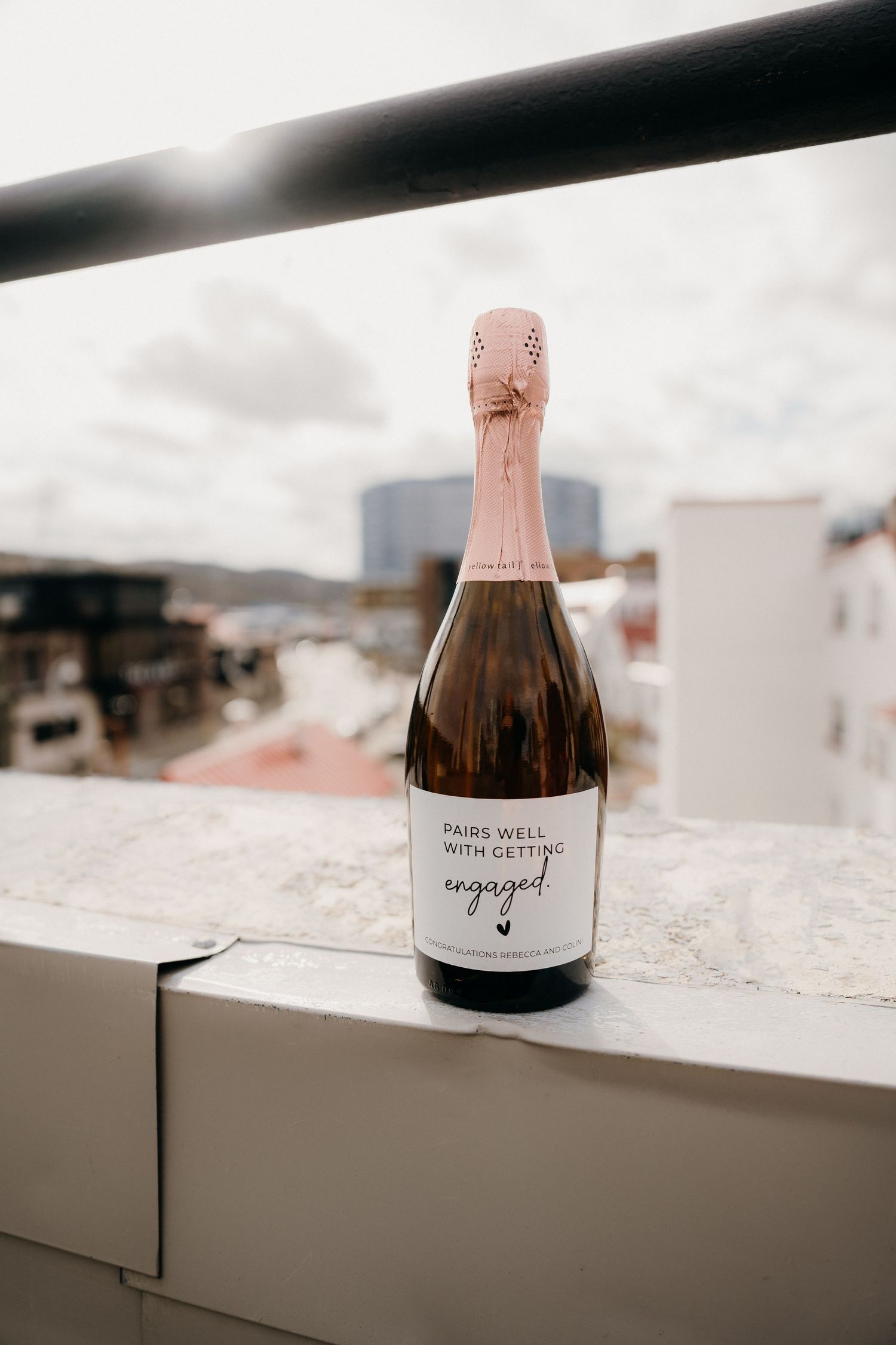 A bottle of rose champagne with 'engaged' text sits on a white ledge overlooking the city.