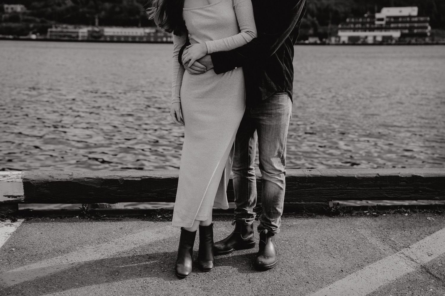 A couple stands together in a romantic moment on a waterfront pier with boats and mountains in the background.