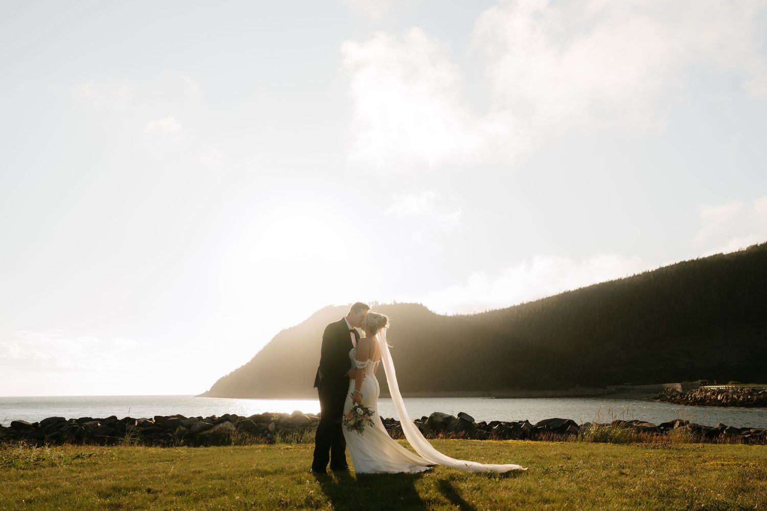 Bride and groom share a romantic kiss at sunset by a scenic lake with rolling green hills in the background.