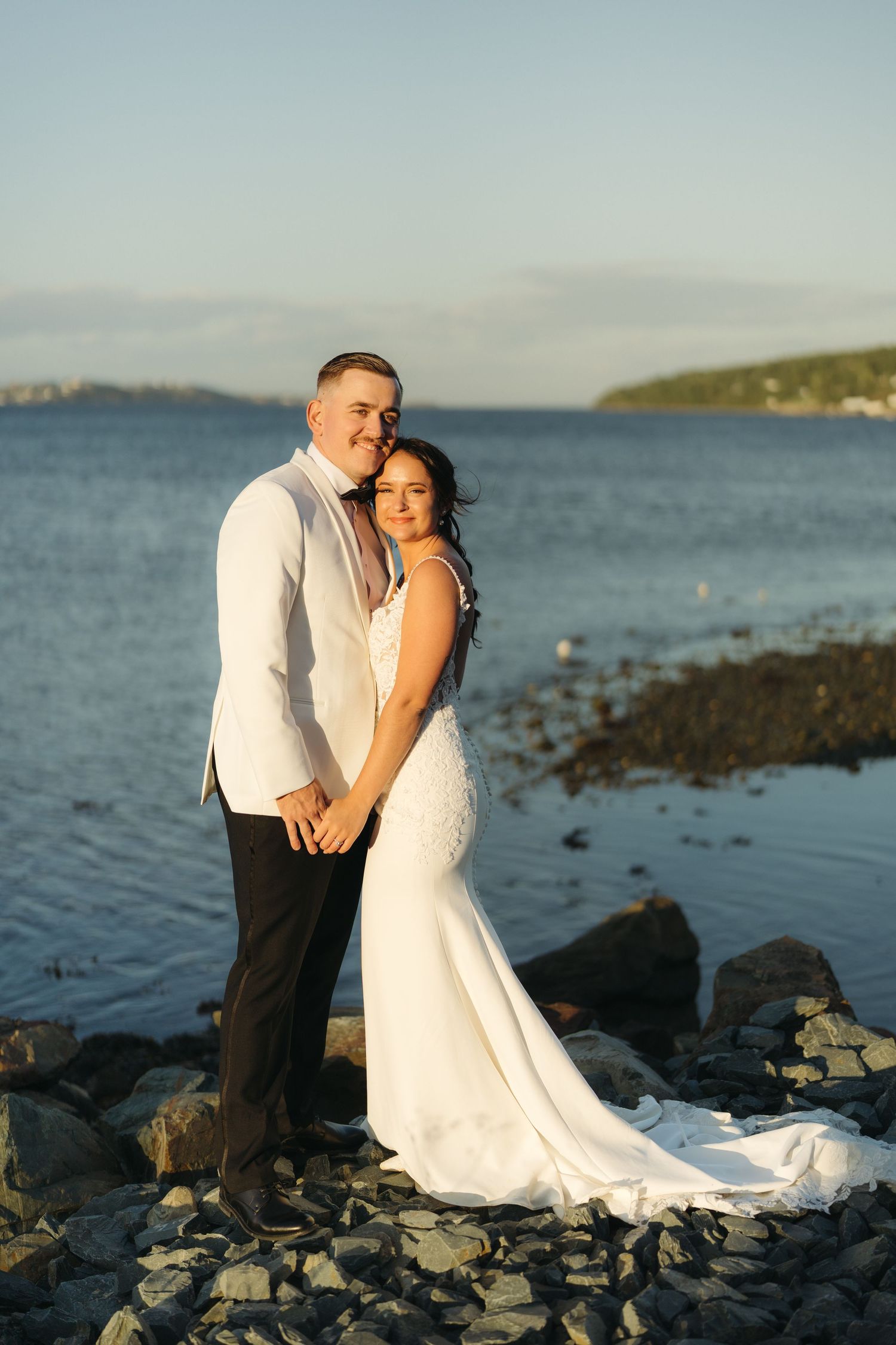 A couple shares an intimate moment on a rocky beach at sunset with the ocean and coastline in the background.