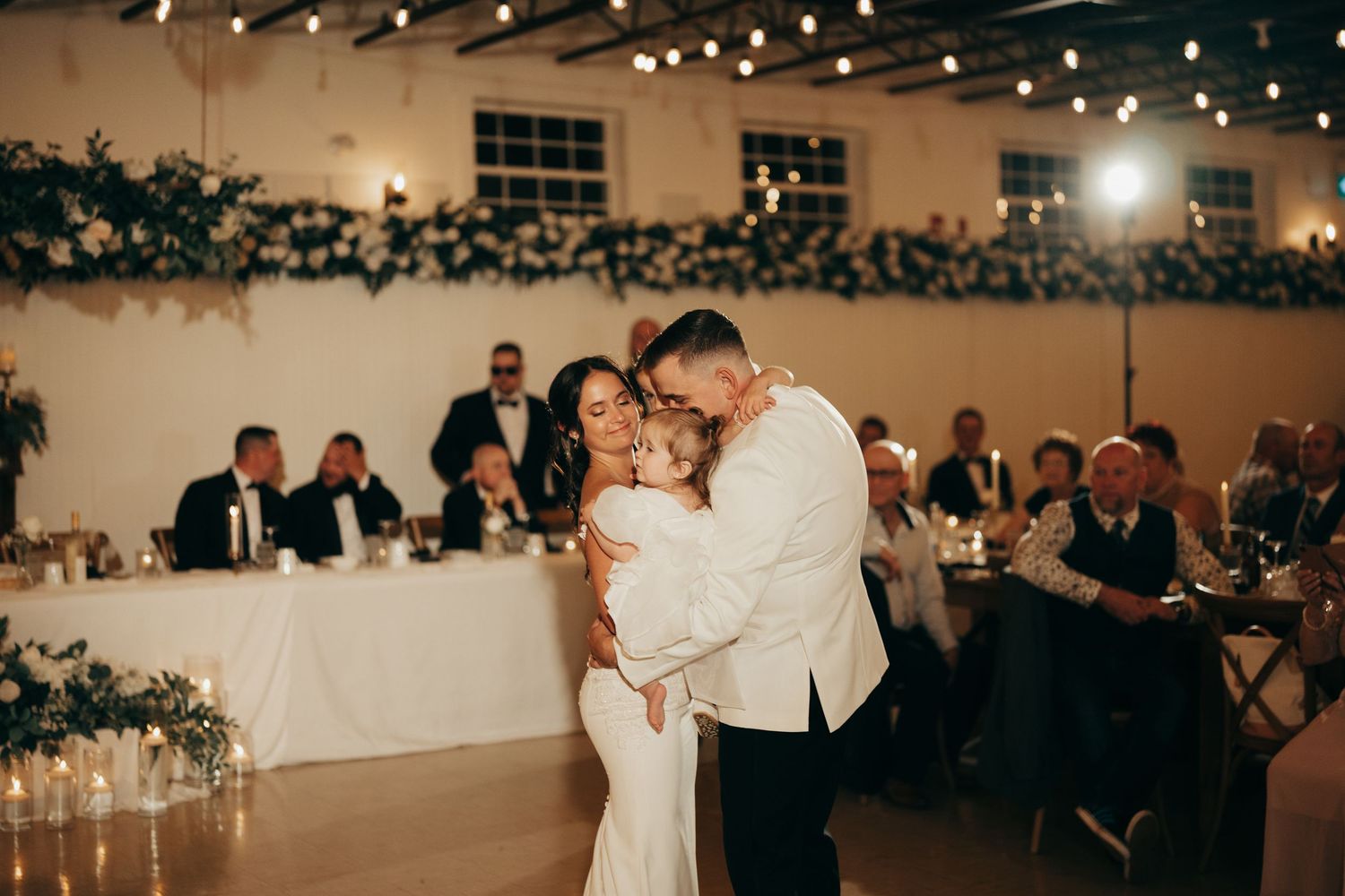 A romantic first dance moment at an elegant wedding reception with twinkling lights and guests watching from their tables.