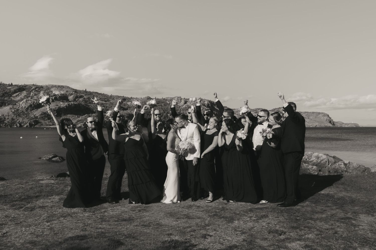 A black and white photograph shows a wedding party celebrating on a scenic cliff overlooking the ocean.