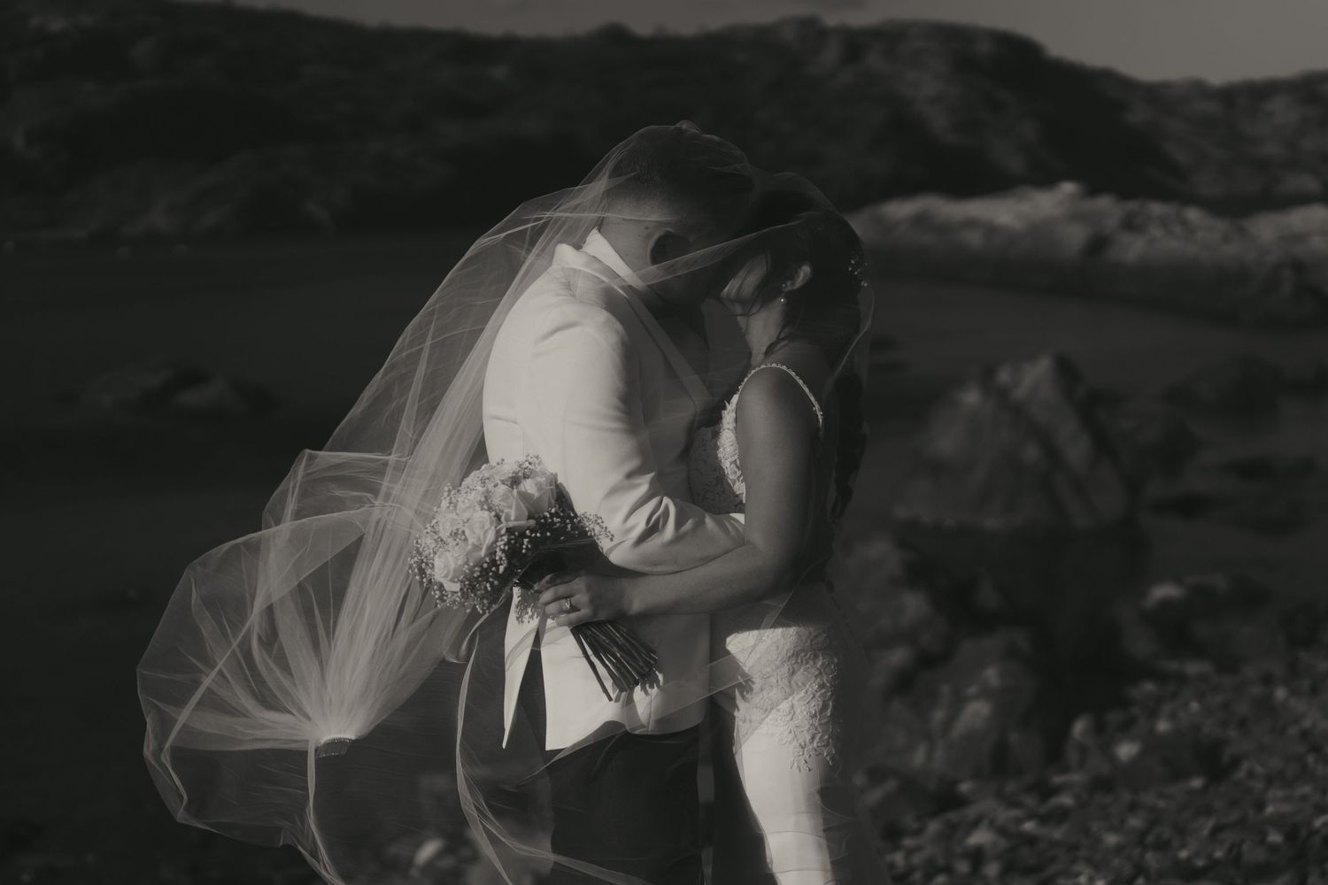 Black and white wedding photograph showing a couple embracing in a romantic moment against a scenic outdoor backdrop.