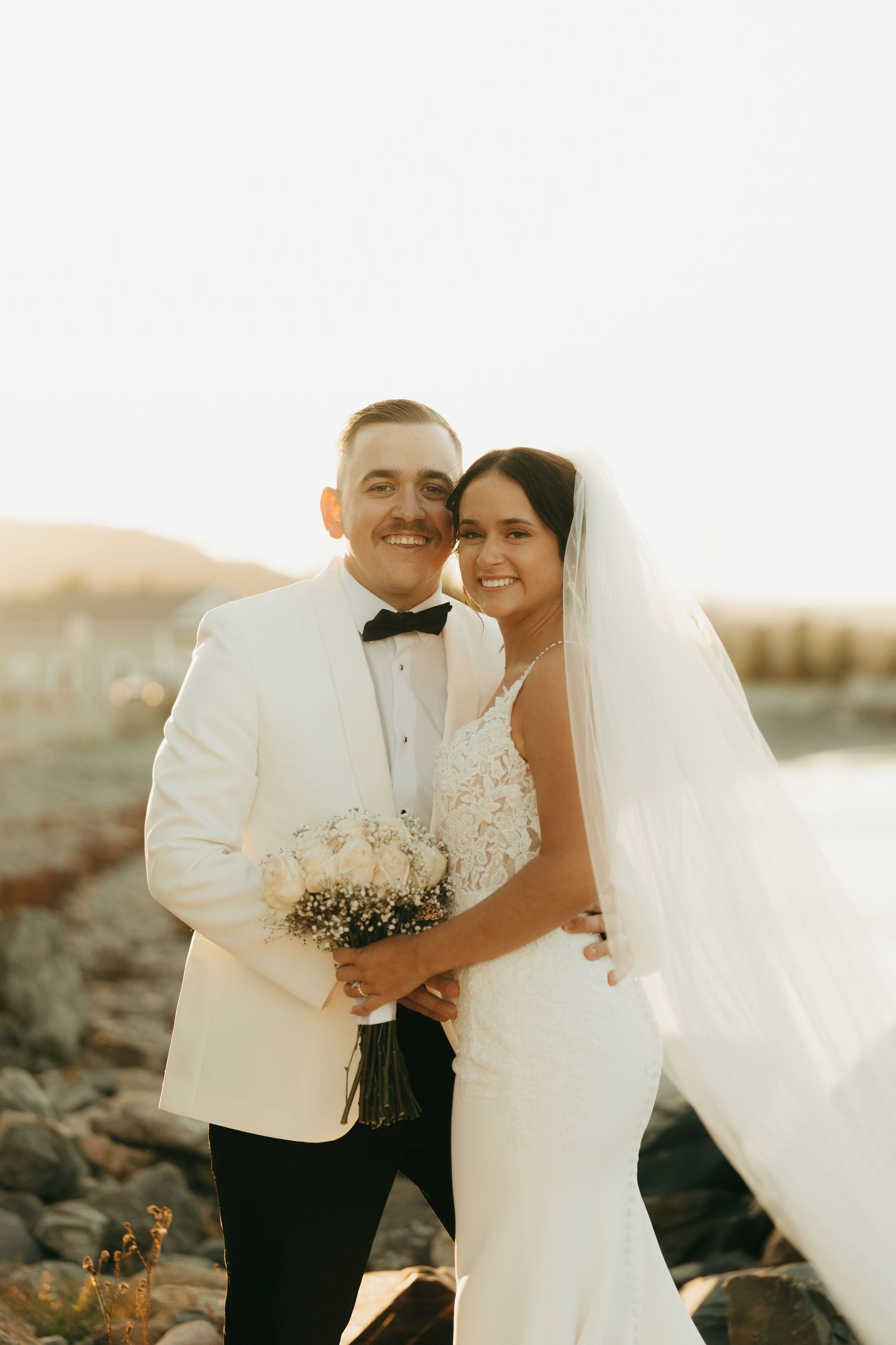 A romantic sunset wedding photo of a couple embracing on a rocky beach in white formal attire.