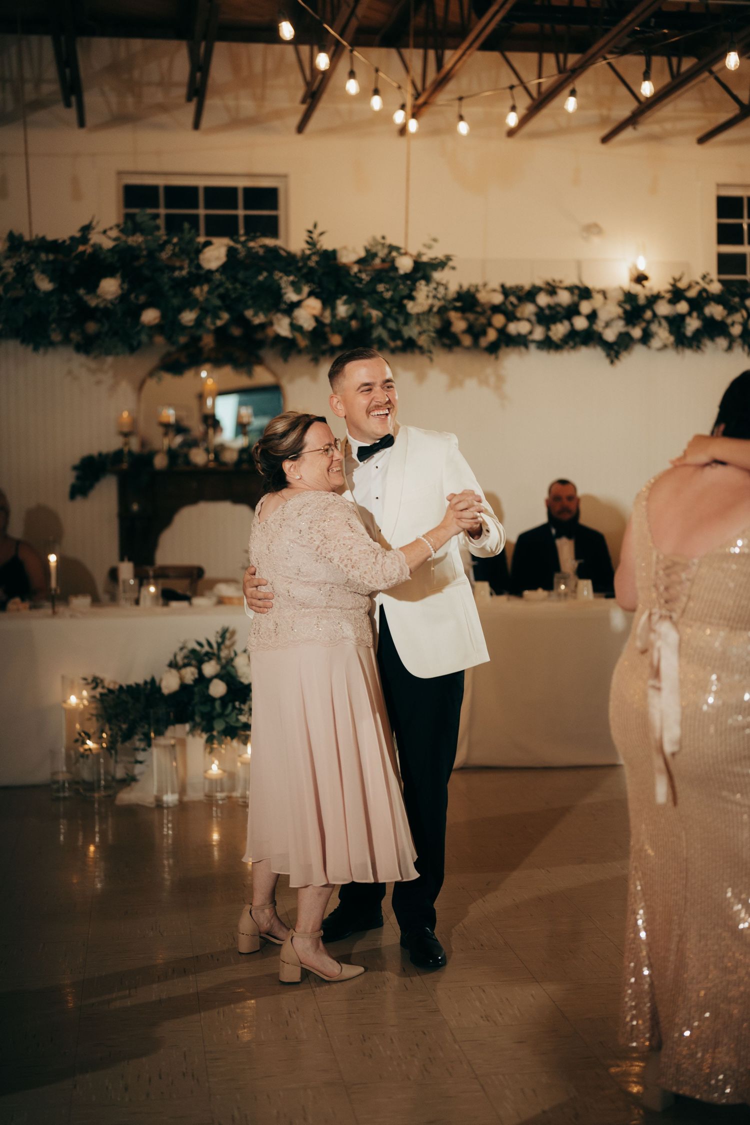 Wedding guests dance and celebrate on an illuminated dance floor at an indoor reception venue with string lights overhead.