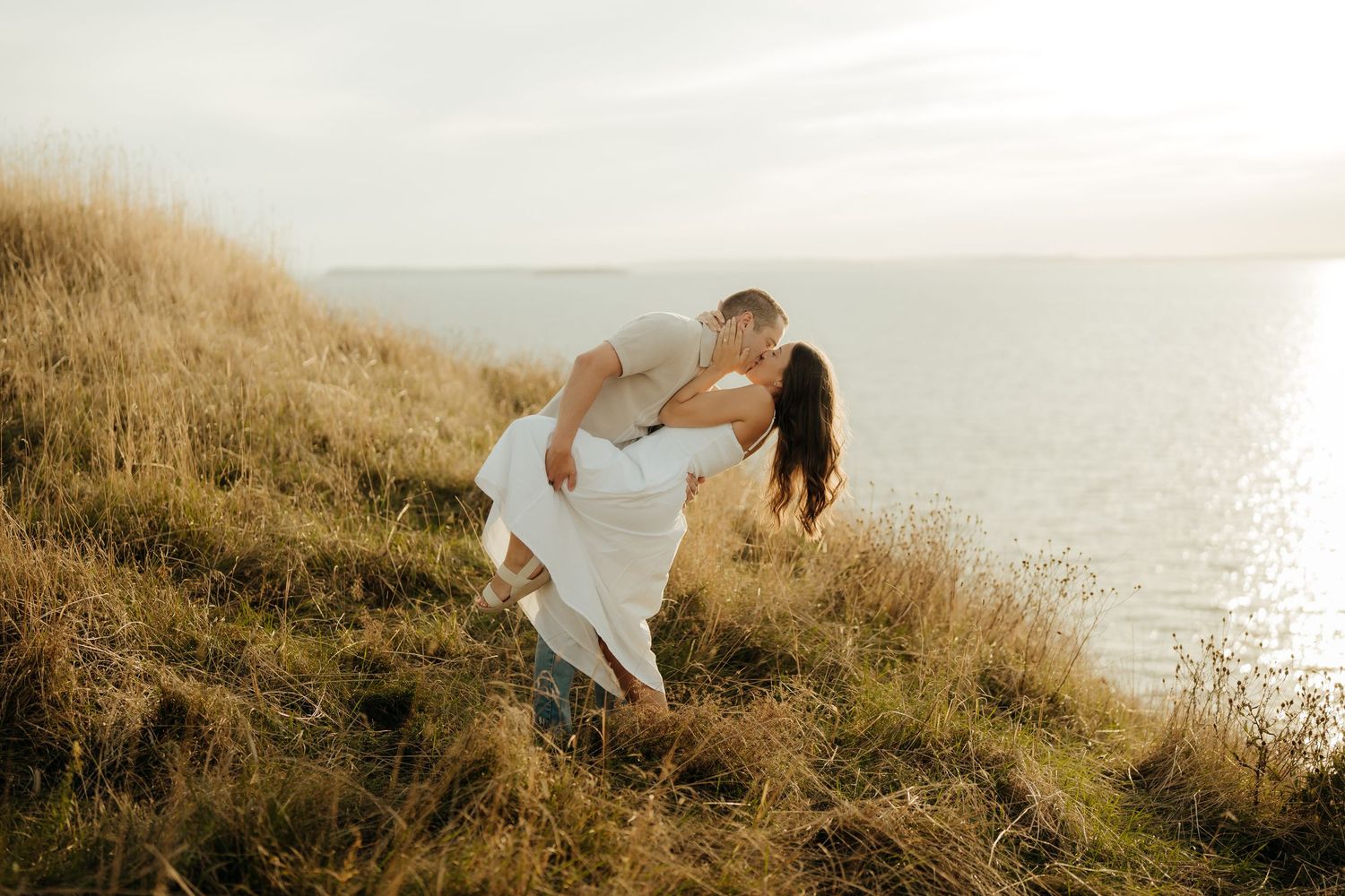 Romantic couple shares tender moments at sunset on a grassy coastal hillside overlooking the ocean.