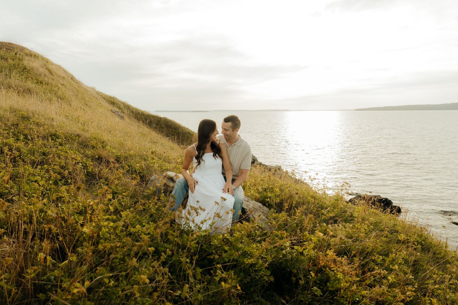 A couple sits on a coastal hillside covered in yellow wildflowers overlooking the ocean at sunset.