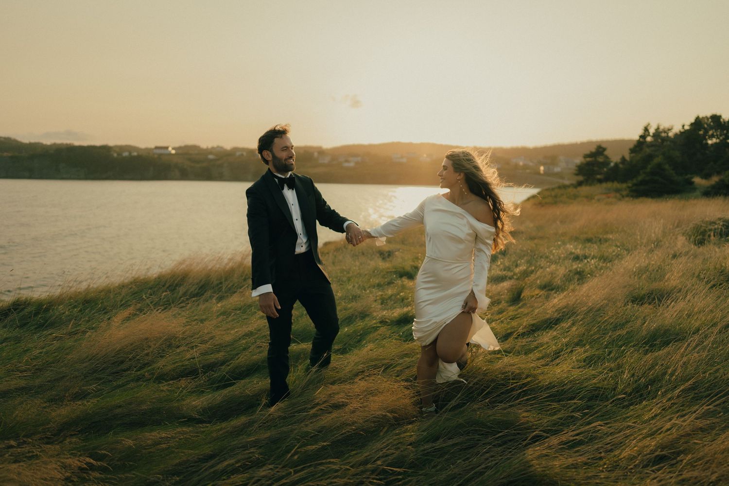 A couple in wedding attire runs through tall grass at sunset beside a serene lake.