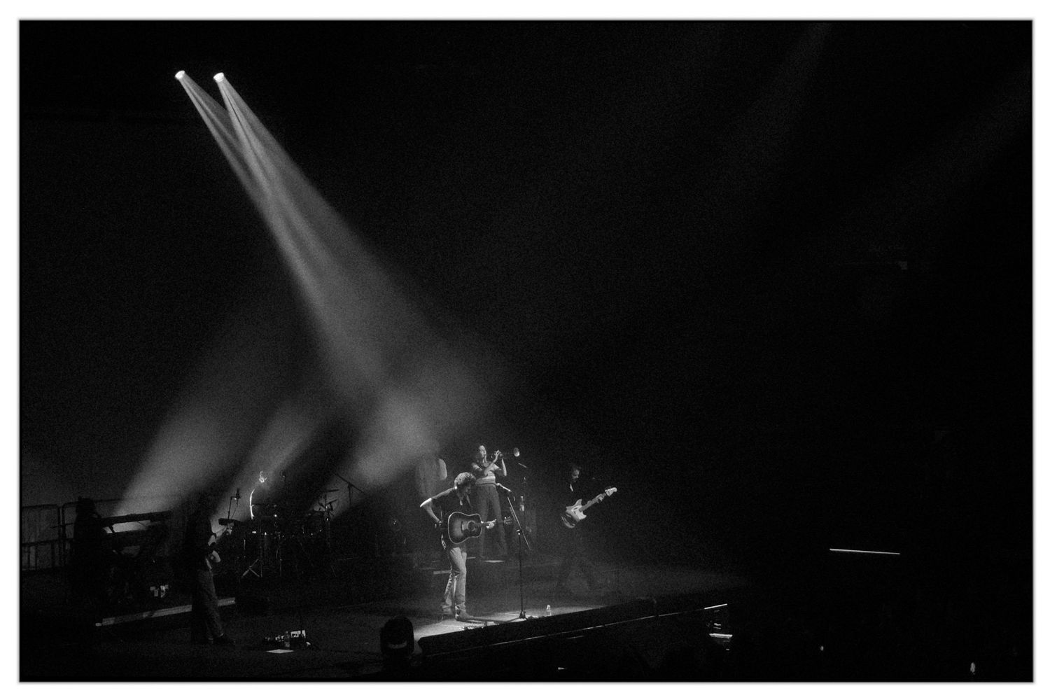 Silhouetted musicians perform on a dark stage with dramatic white spotlights illuminating their performance.