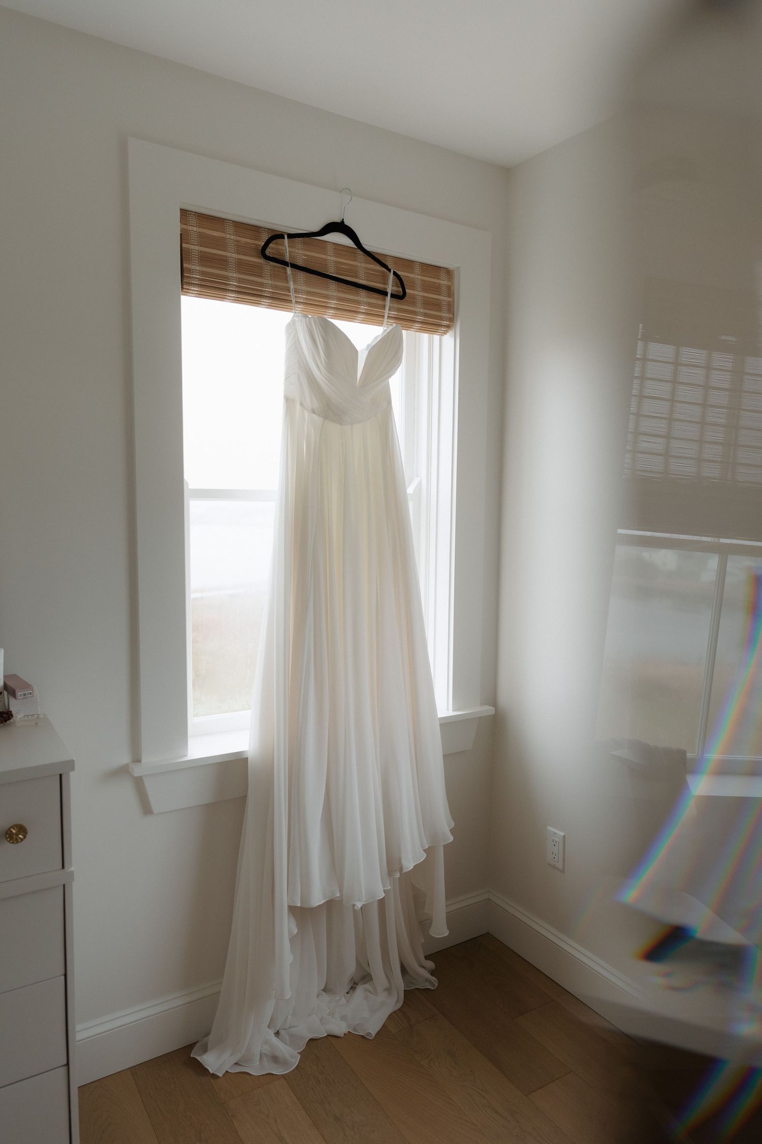 A white wedding dress hangs gracefully from a hanger near a bright window in a minimalist room.