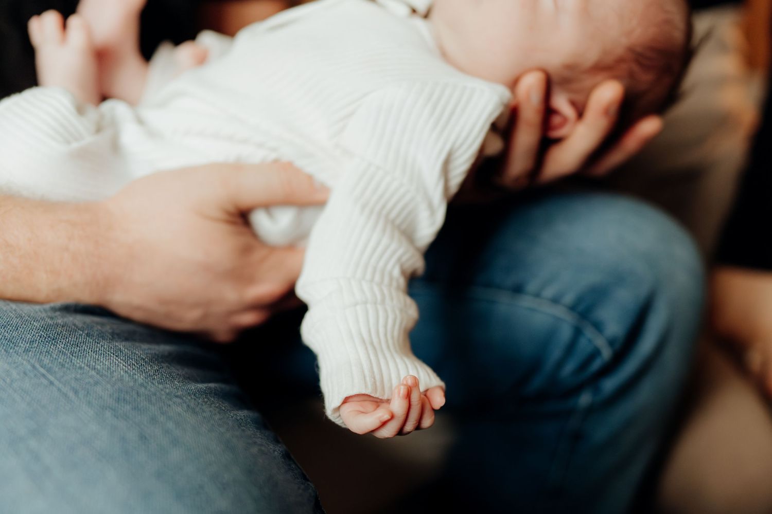 Close-up view of tiny baby feet and hands being held gently against denim-clad legs.