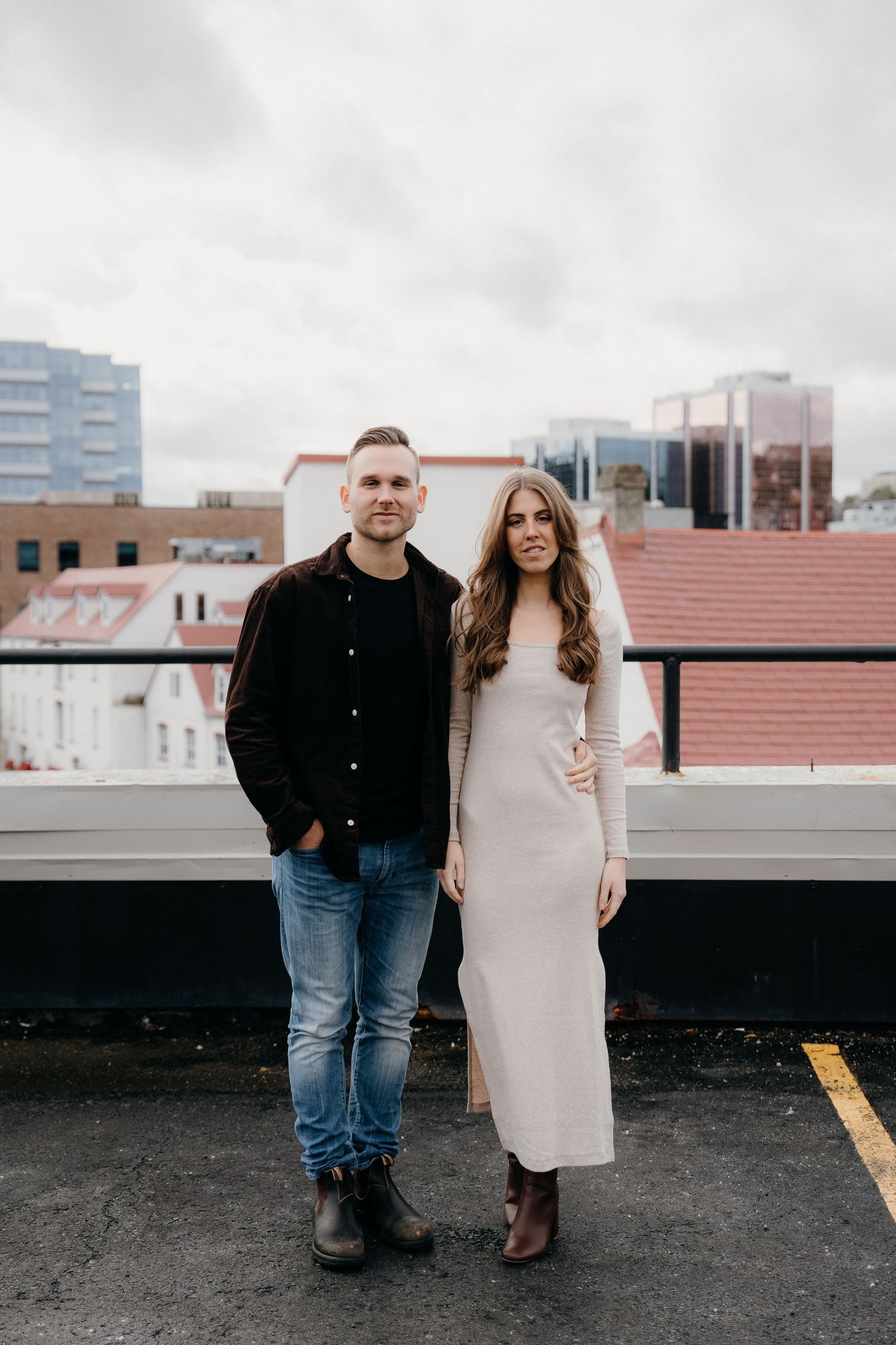 Couple in casual attire standing together on empty rooftop parking area with city views.