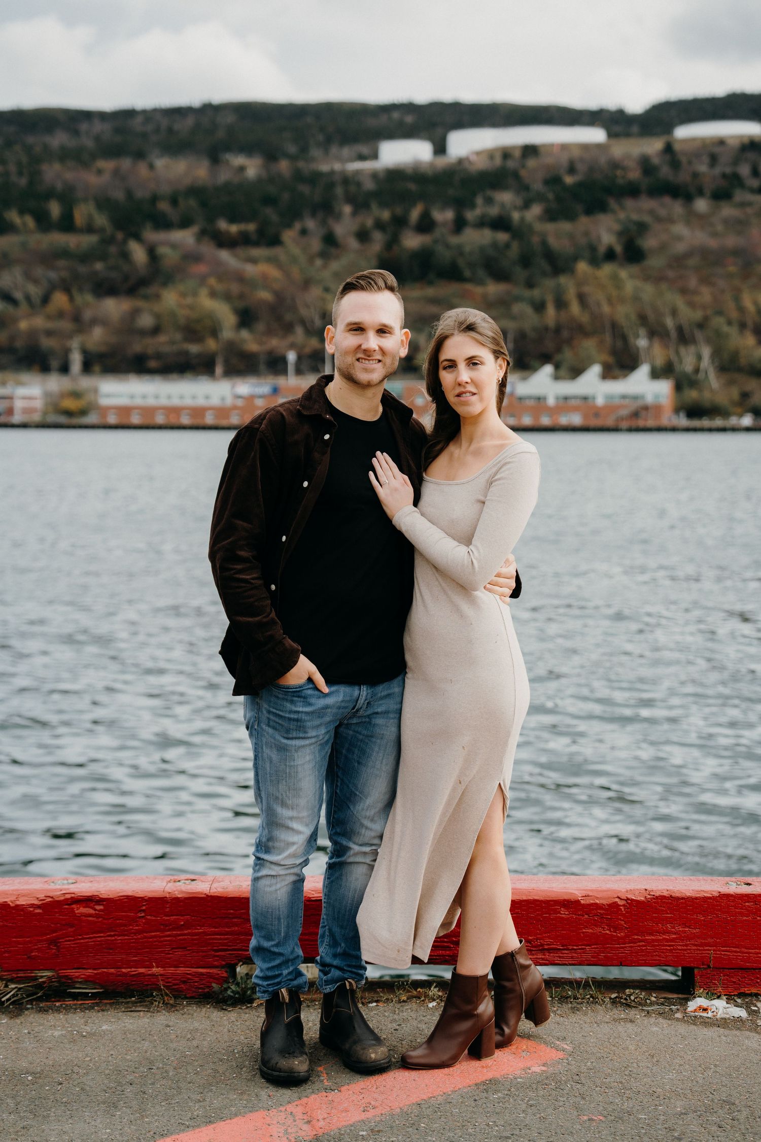 Engagement photo of couple standing on red-painted dock with water and hills behind them.
