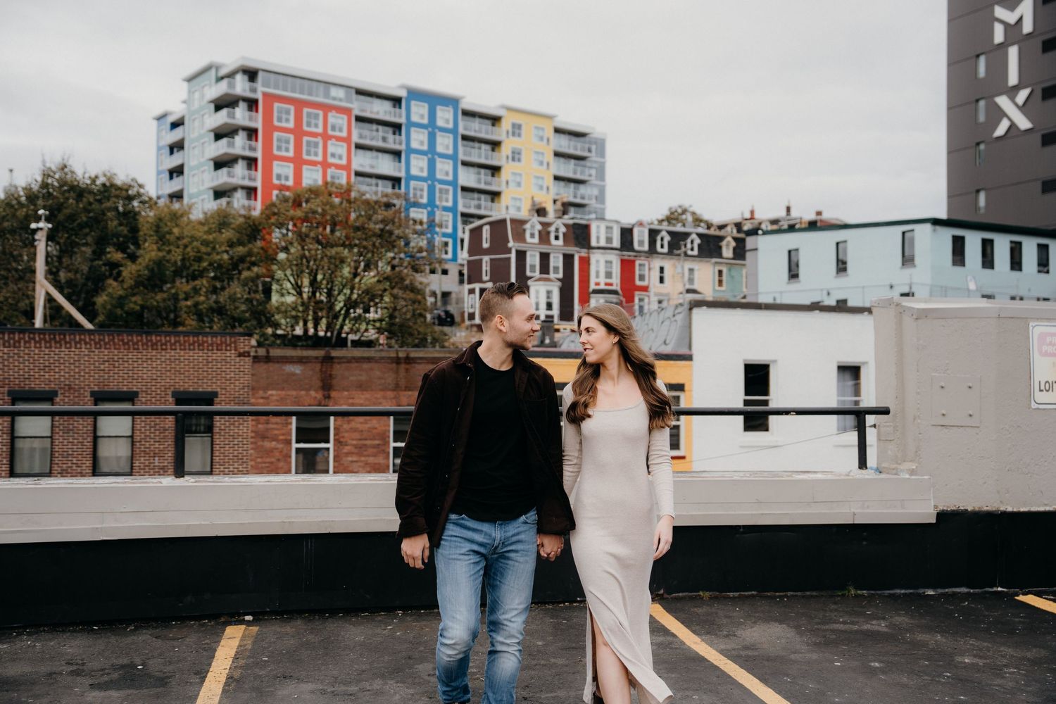 Couple embraces in an urban setting with colorful buildings and a cloudy sky in the background.