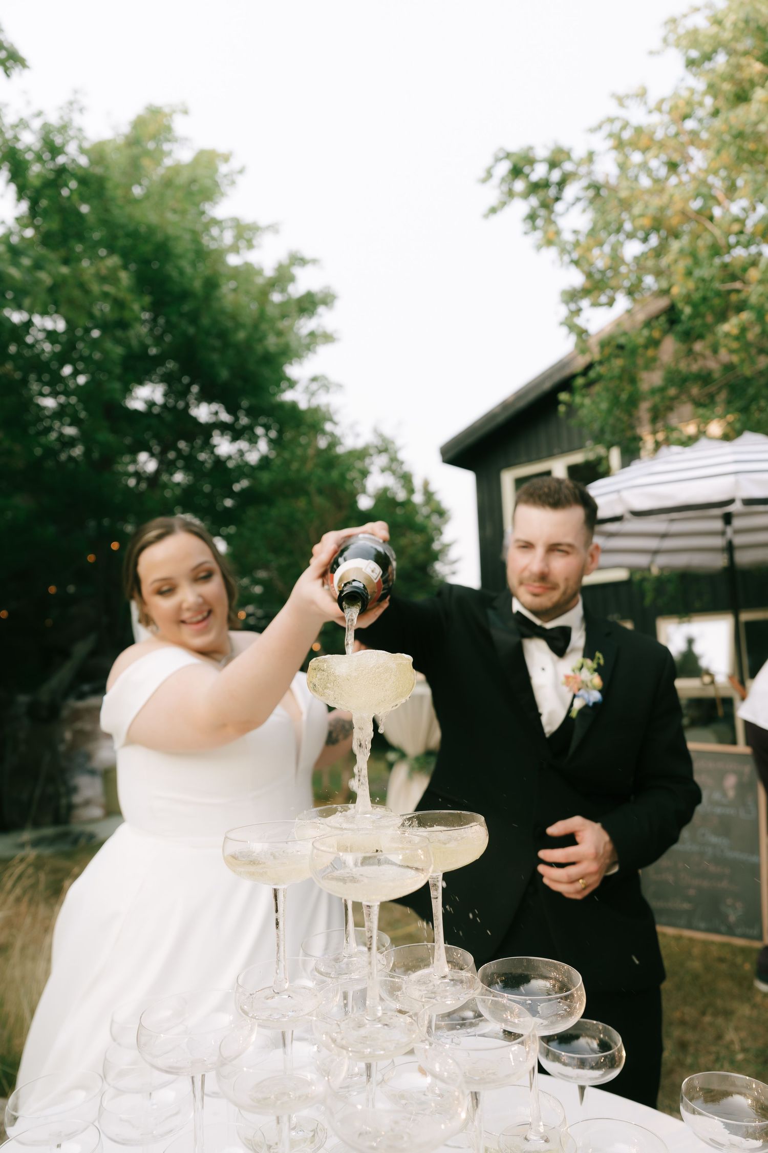 Newlyweds carefully pour champagne into an elaborate tower of coupe glasses at their reception.